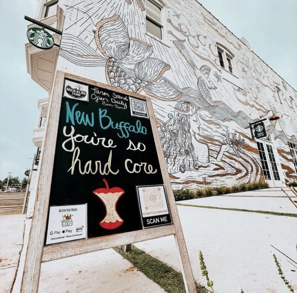 A sidewalk chalkboard sign outside a building with a large mural. The sign advertises a new Buffalo offering, with open daily hours from 8 am to 3 pm, and has QR codes for scanning. The mural features a woman holding a torch and is decorated with lar