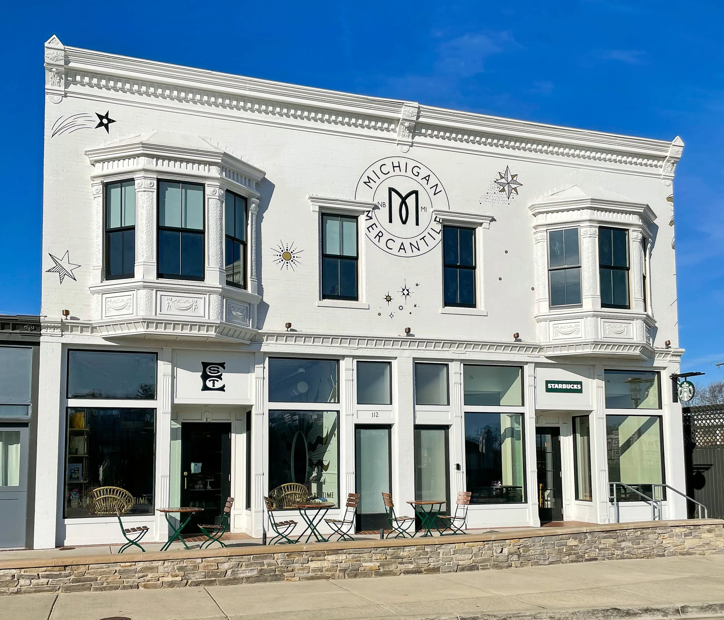White three-story building with large bay windows, outdoor seating on the sidewalk, and a sign indicating a Starbucks cafe. Decorative stars and celestial designs on the facade. Clear blue sky.