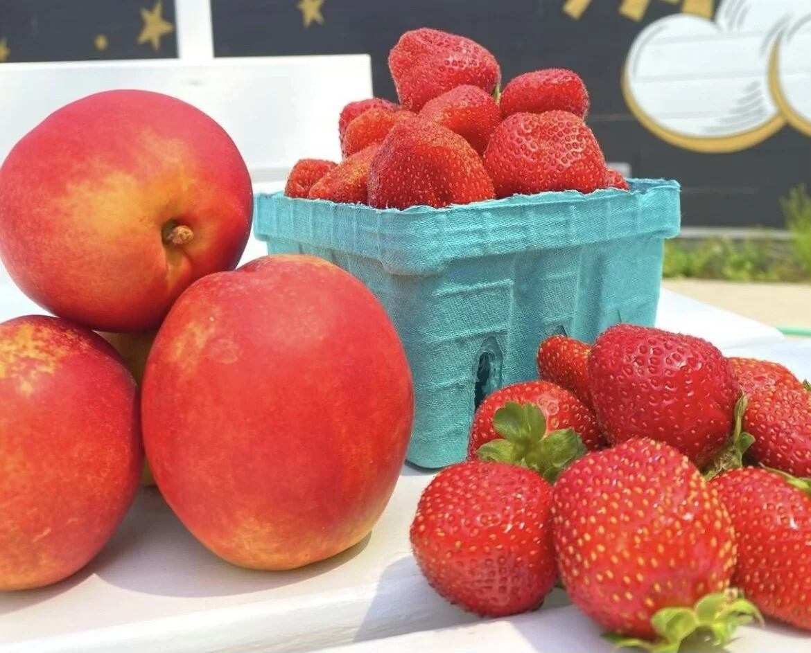 Three large apples and a bunch of strawberries on a white surface, with a basket filled with more strawberries in the background.