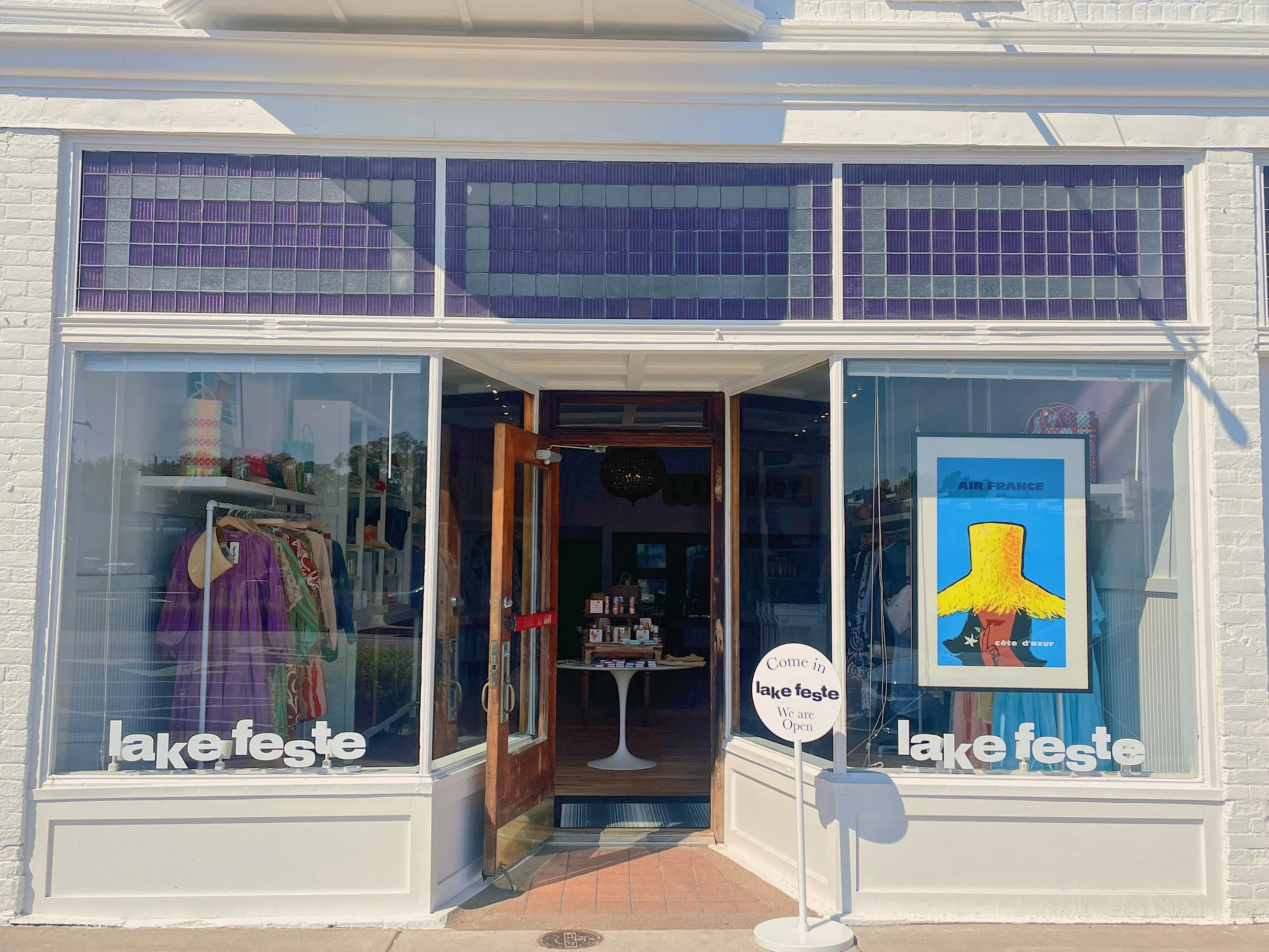 Exterior of a clothing boutique with large glass windows, displaying colorful clothes inside. A sign near the entrance reads 'Come in lake feste. We are Open.' A large framed picture of a hat with a yellow brim, and a poster with the same hat, is visible in the window.