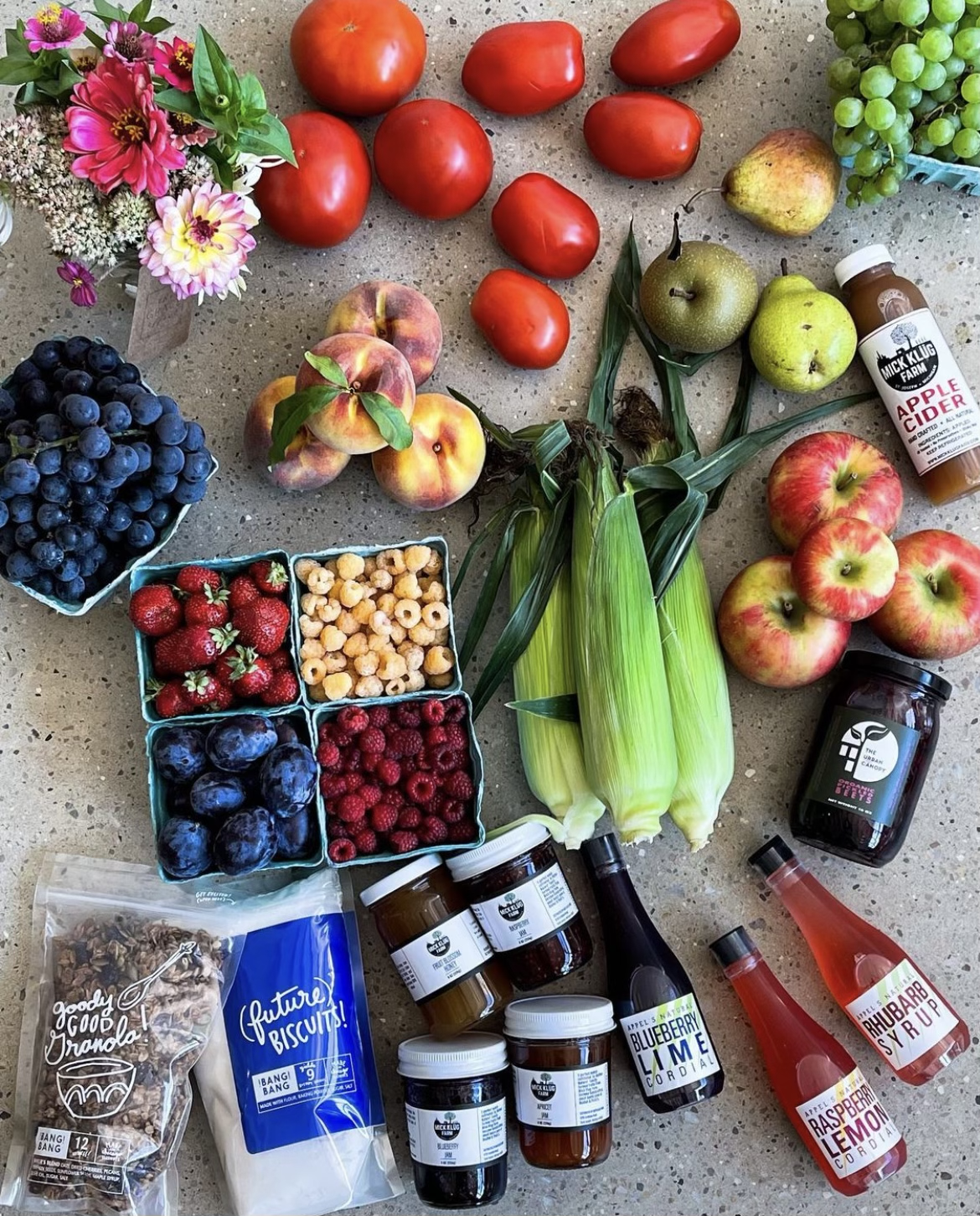 Various fresh fruits including tomatoes, peaches, pears, apples, strawberries, raspberries, blueberries, and grapes, along with bottles of fruit syrups, apple cider vinegar, honey, a bouquet of flowers, and jars of jams or preserves, spread out on a 