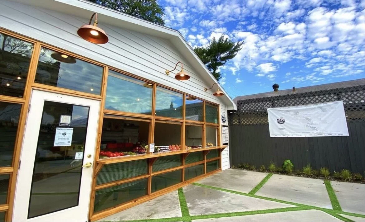 Exterior view of a small shop with a wooden framed window display and a white door. Items such as strawberries and other produce are visible inside. The sky is partly cloudy, and the pavement outside has green painted lines.