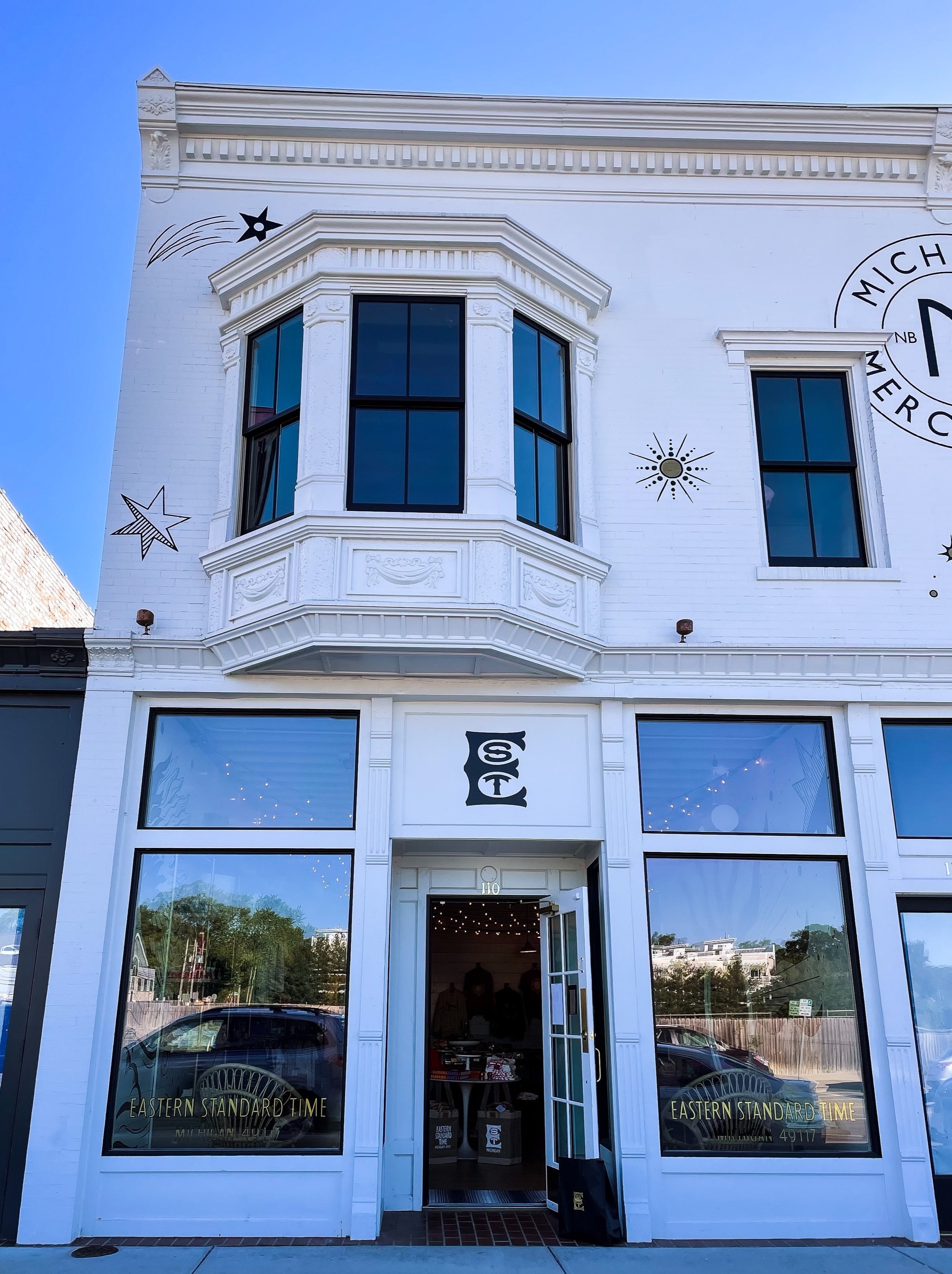 White two-story building with large front windows, decorative molding, and mural art including stars and a circular logo on the upper right. The entrance has open glass doors, and signs on the windows read "Eastern Standard Time" and the address number 110.