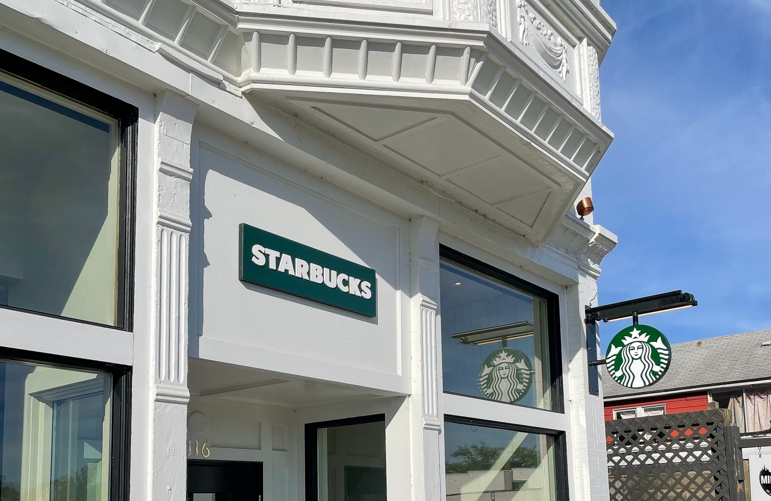 Exterior of a Starbucks store with a green and white logo sign and a shorter green sign reading ‘Starbucks’ on a white building, with large windows reflecting the blue sky.