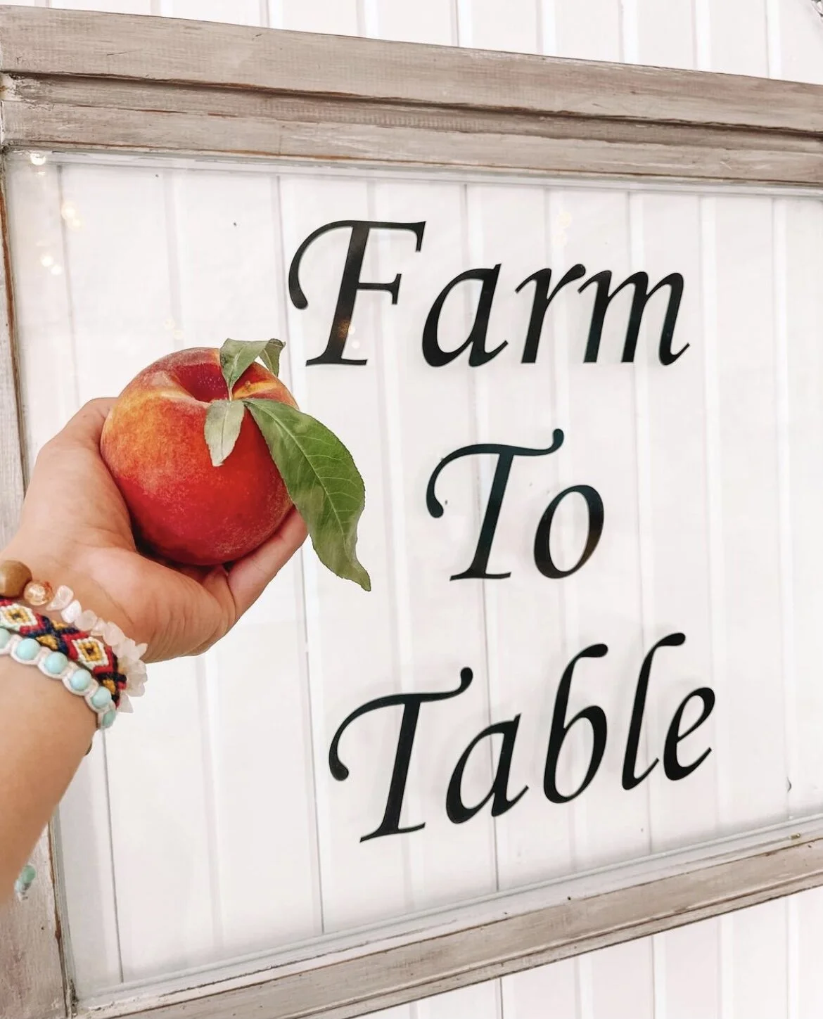 A person holding a red apple with green leaves in front of a sign that says 'Farm To Table'
