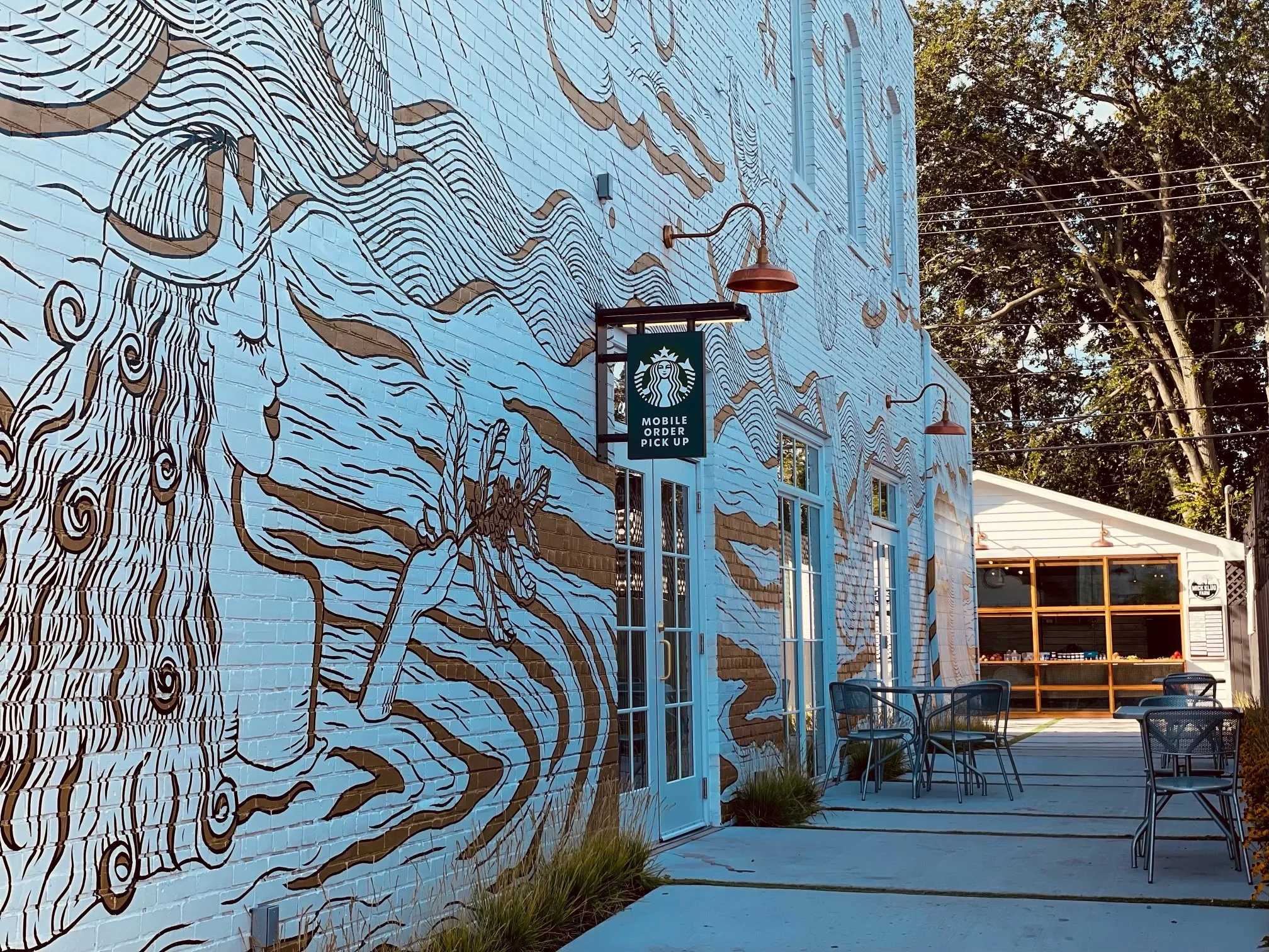 Exterior of a building with large mural art featuring a mermaid and a lobster, painted on a brick wall, with outdoor seating and a Starbucks sign indicating mobile order pickup.