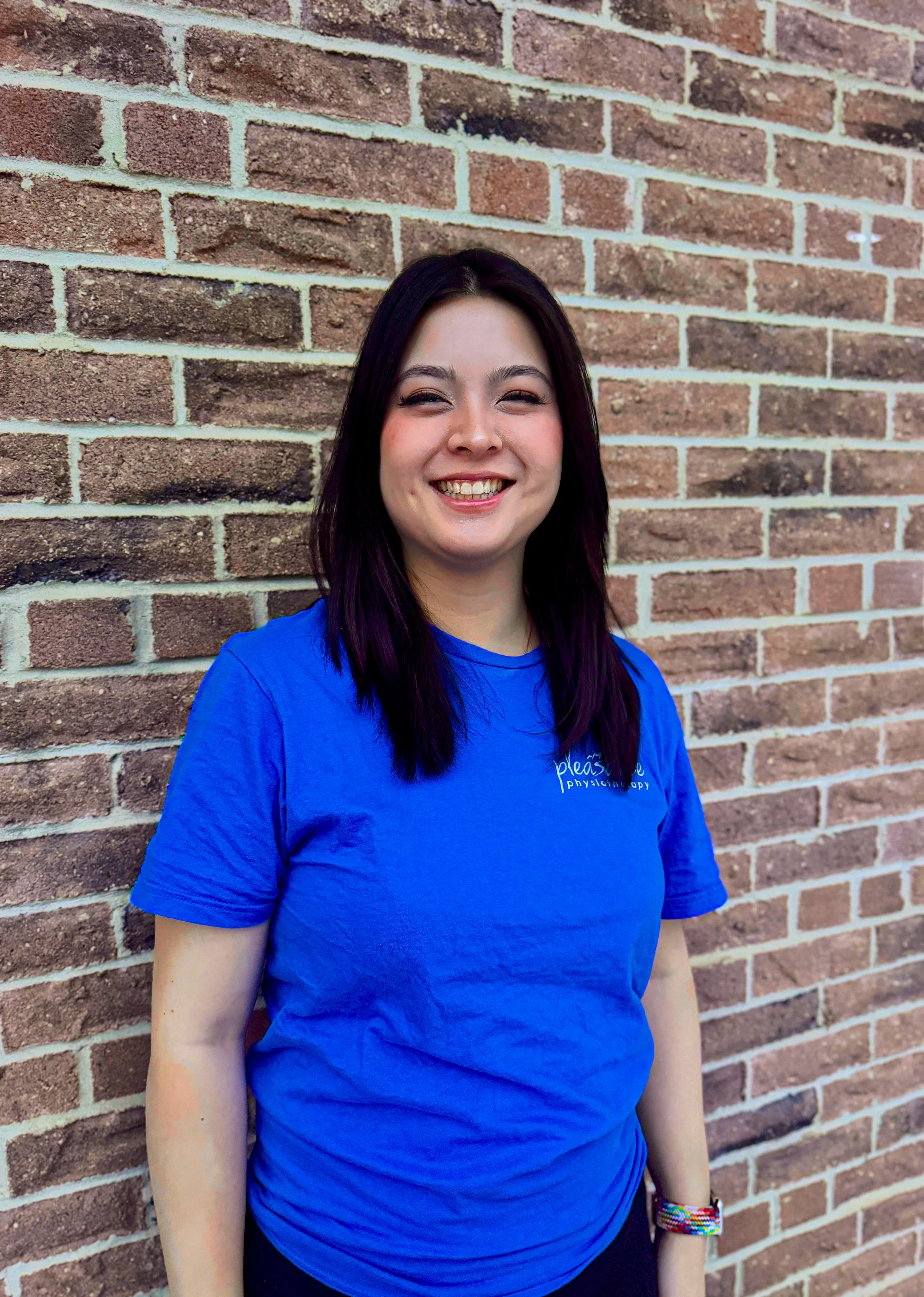 Portrait of a young woman with short dark hair, smiling, wearing a black top, a silver necklace with a four-leaf clover pendant, and a green beaded bracelet, standing against a light-colored wall.