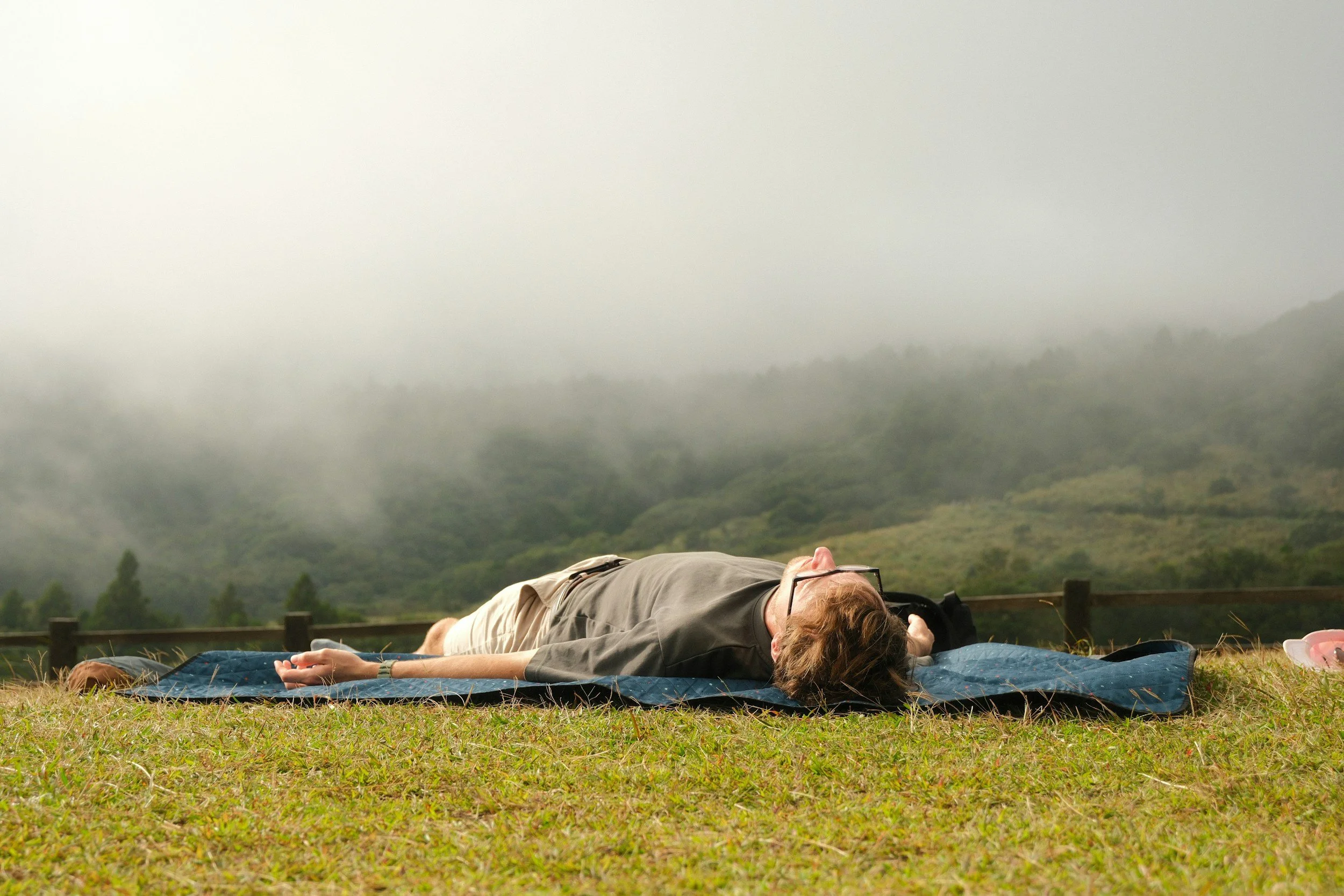 A person with medium-length, honey-colored, slightly tousled hair lays outdoors on a dark blue yoga mat placed in the grass on a foggy morning in the countryside. The relaxing person wears glasses and a wristwatch.