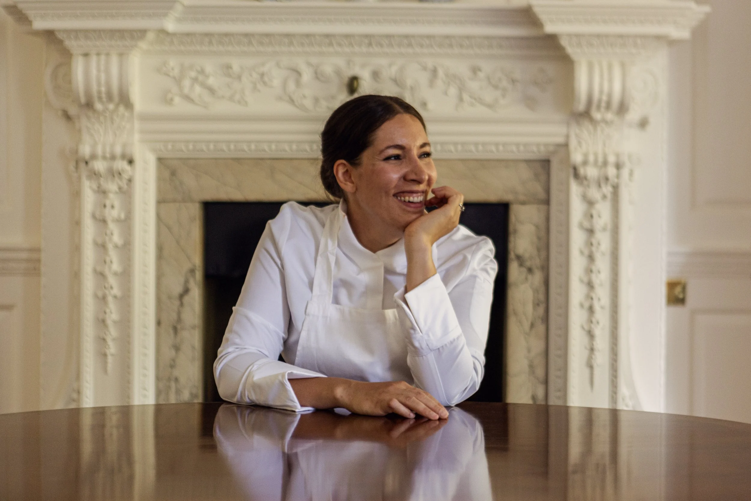Sarah, in a white chef's coat smiling, resting her chin on her hand, sitting at a polished wooden table in a room with intricate white marble fireplace and cream-colored walls.