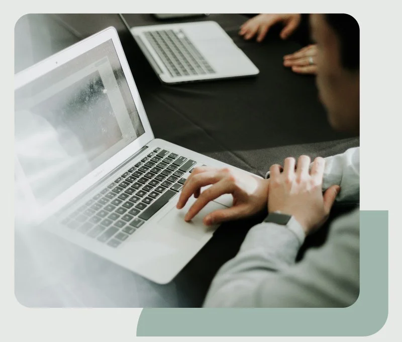 Person working on a laptop at a meeting with another person, with additional laptop on black table.