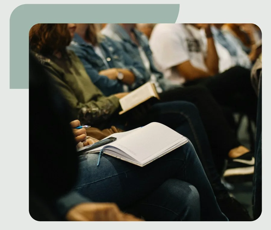 People sitting in a conference or classroom setting, some taking notes or reading.