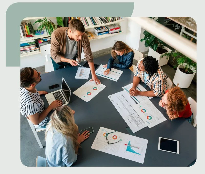 Group of six diverse professionals in a meeting room with laptops and documents, discussing business charts and graphs.