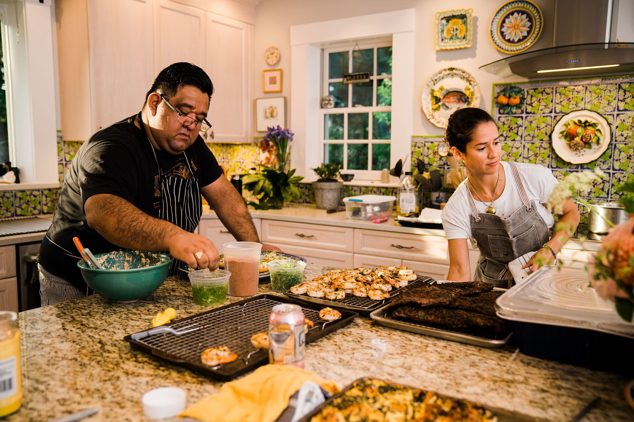 Chef Fernanda Tapi doing a cooking class in a colorful kitchen with a man dressed in black top and striped apron.