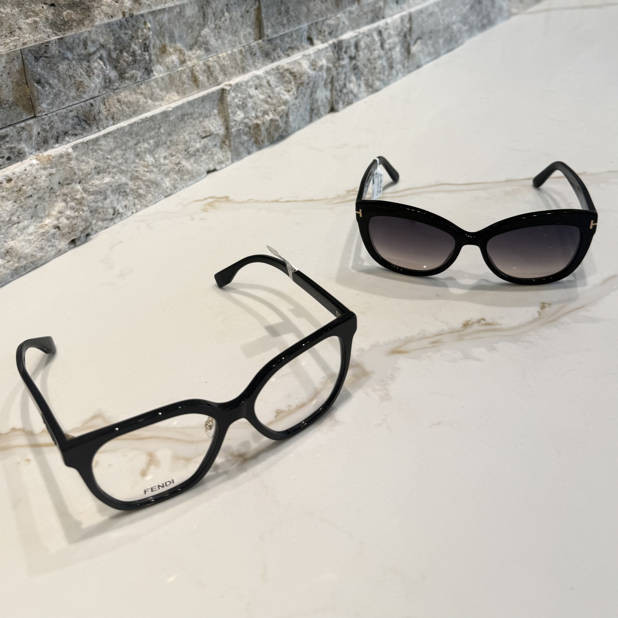 A pair of clear ophthalmic Fendi frames next to a pair of polarized Tom Ford sunglasses on a quartz counter with light colored stone in the background