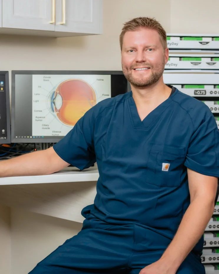 Dr Dornstauder wearing scrubs and sitting in his exam room in front of a computer with an eye diagram and there are contact lens trays behind him