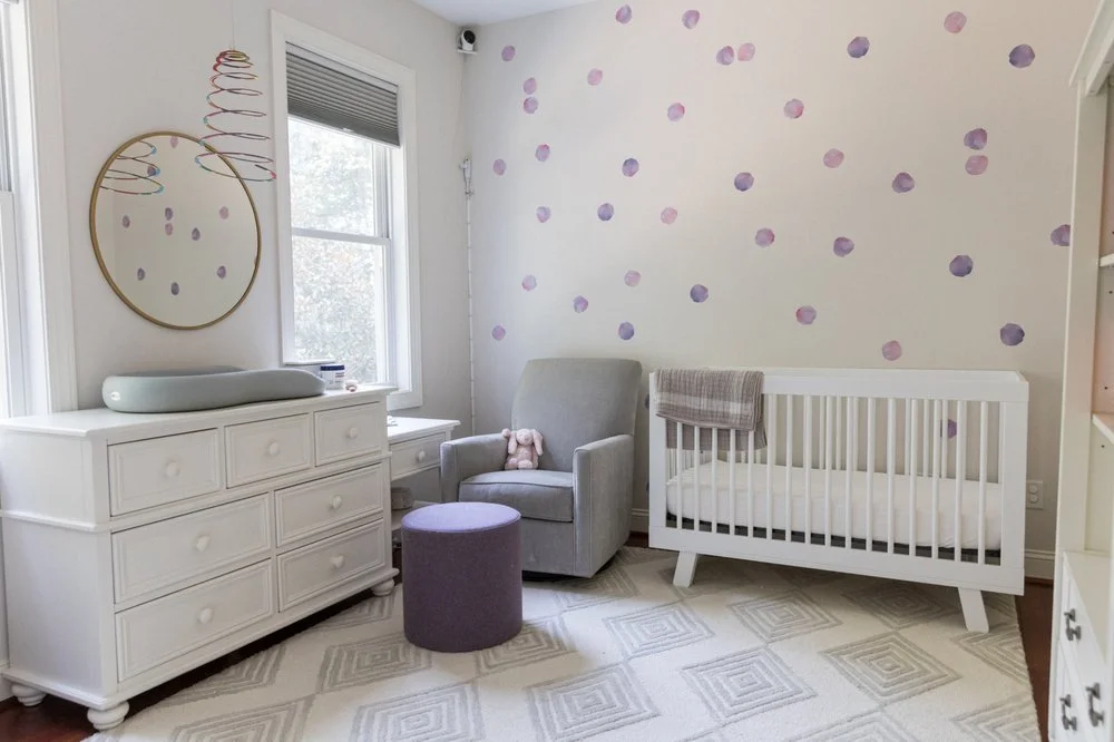Well-organized children's nursery with white crib, dresser, wall art, rocking chair, colorful ottoman, gray/white rug, and more.  