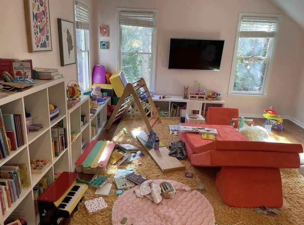 Before. A designed children's playroom with large windows, colorful toys, a small slide, a shaggy rug, plush toys, wall-mounted animal heads, and a wall-mounted TV.