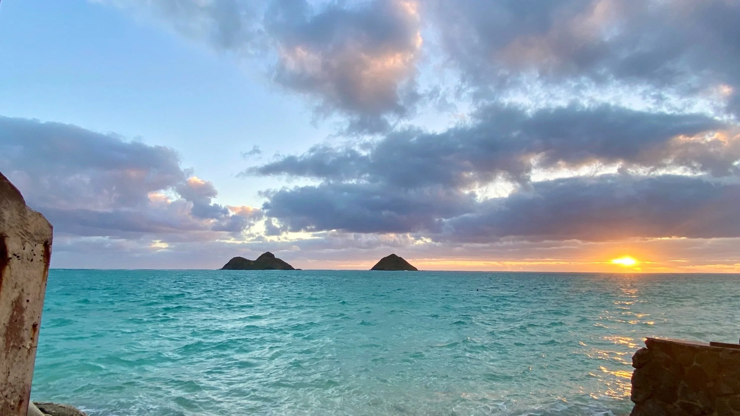 Sunrise over a tropical ocean with two small islands on the horizon, partly cloudy sky, and a rocky foreground.