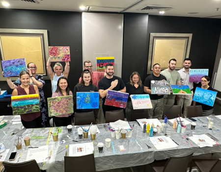 Group of people holding up colorful paintings in a room with a long table covered in art supplies and paper.