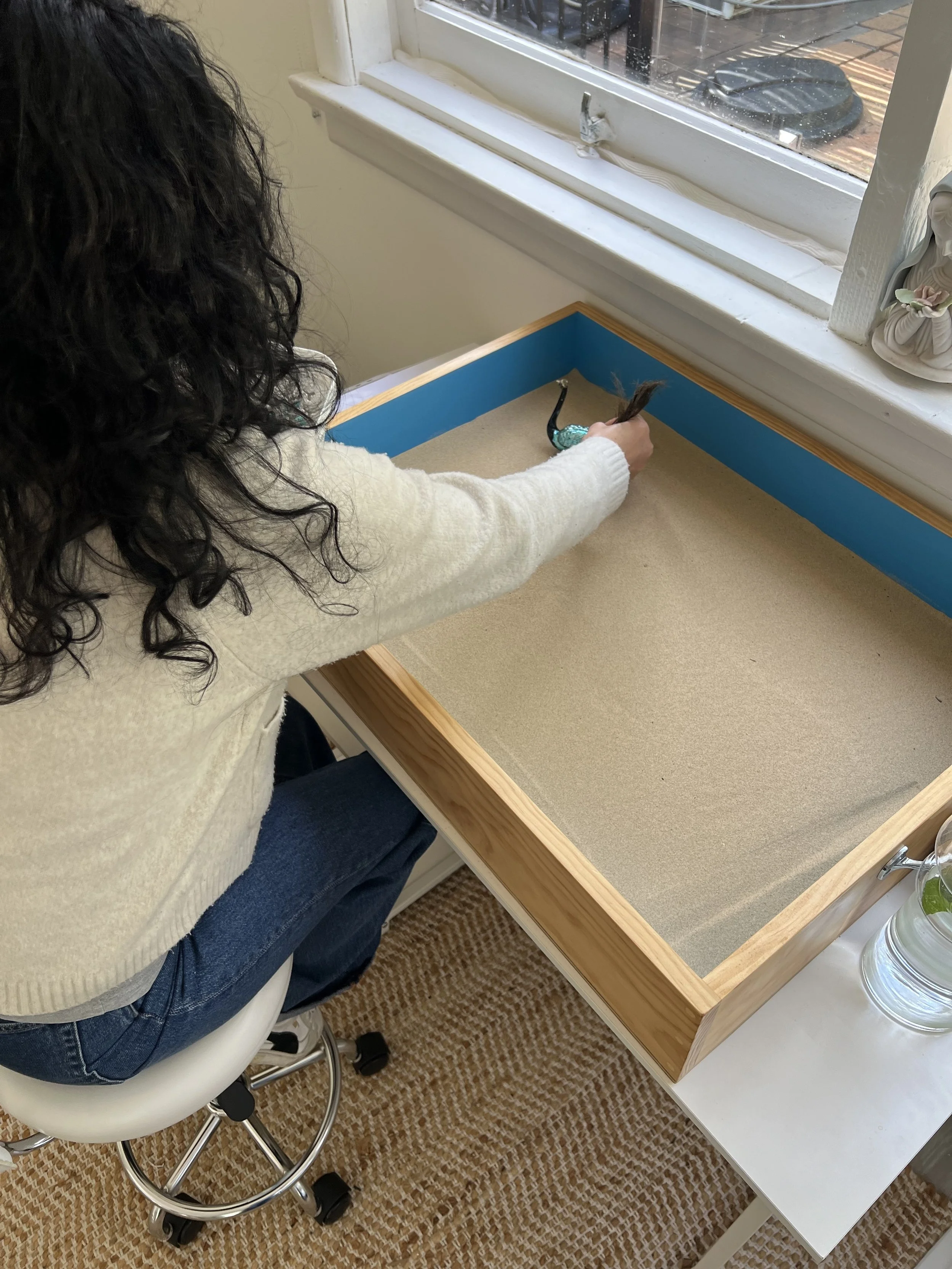A woman with long, curly black hair wearing a cream sweater and jeans sitting on a white rolling stool at a wooden table with a sandbox base, placing a small decorative swan figurine inside. There is a window with a view outside and a small decorativ