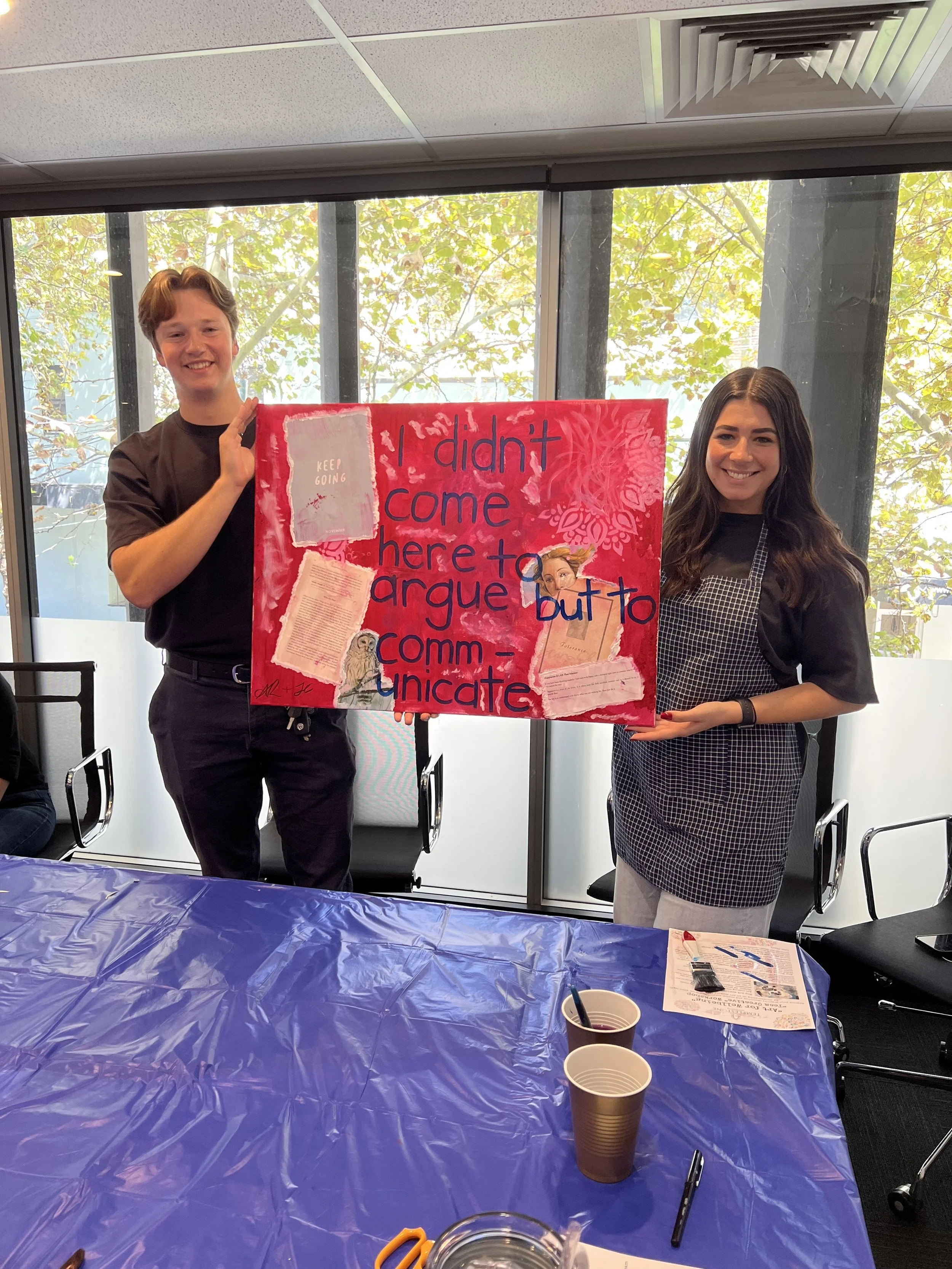 Holistic Art Therapy workshop for corporate wellbeing. Two employees displaying their collaborative art piece. The poster reads: "I didn't come here to argue but to communicate" and includes various images and decorations.