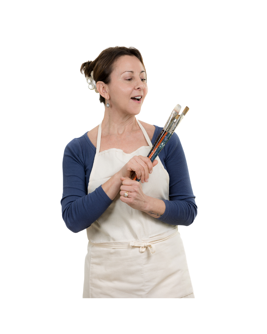 A woman in a white apron holding paintbrushes, looking at them with interest.