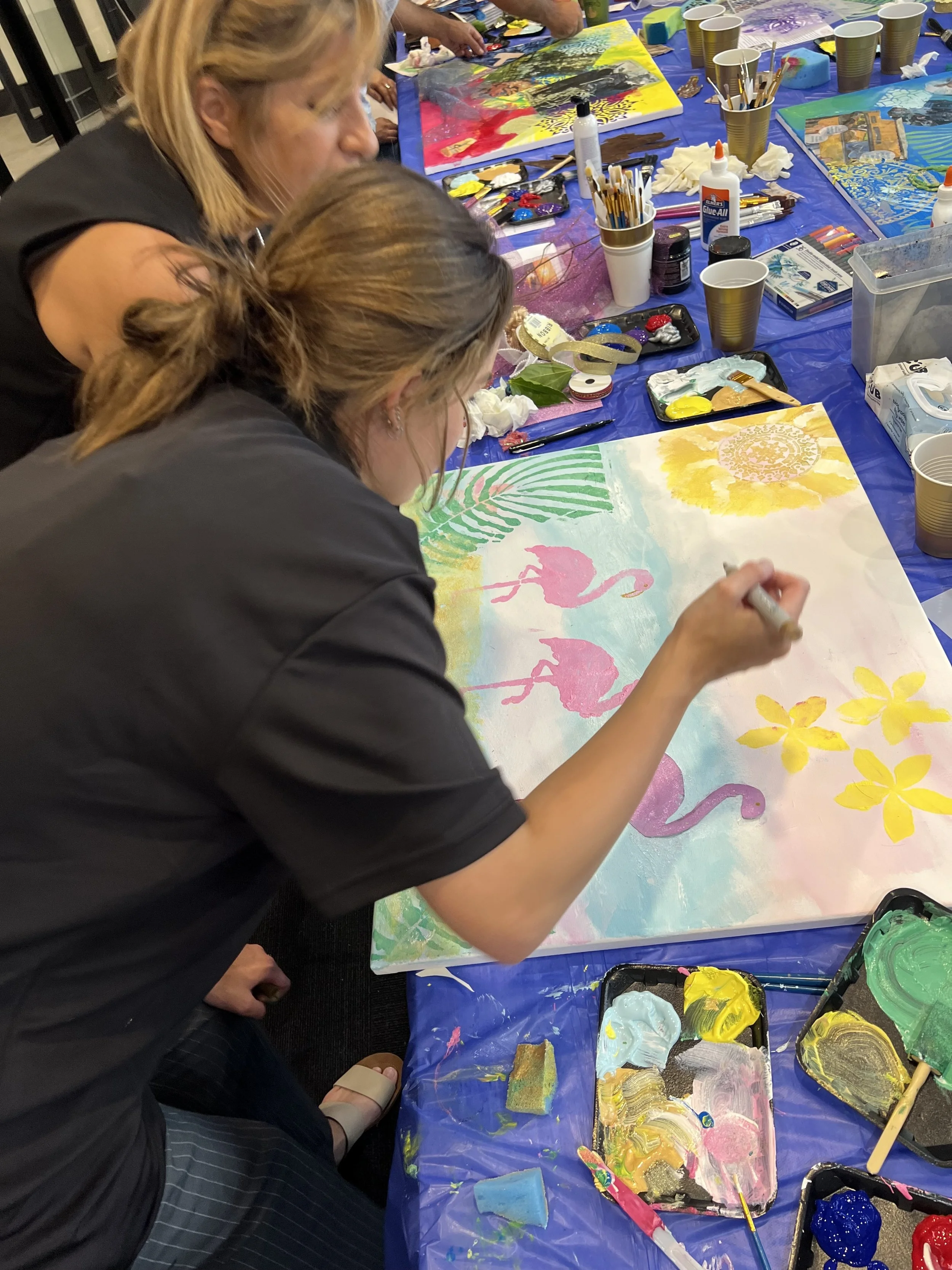People painting a tropical-themed artwork with pink flamingos, yellow flowers, green leaves, and a yellow sun on a large canvas at an art party.