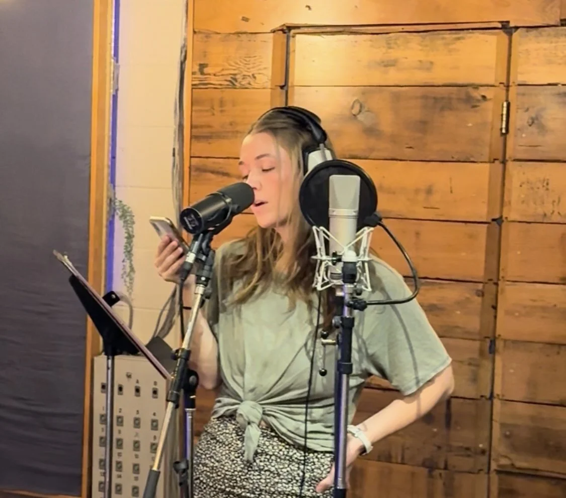 Greta Craig recording the fiction podcast, "Violet's Shadow: A Murder Mystery"  in a music studio with wooden walls, using a pop filter, wearing headphones, holding a phone, and standing in front of a microphone.