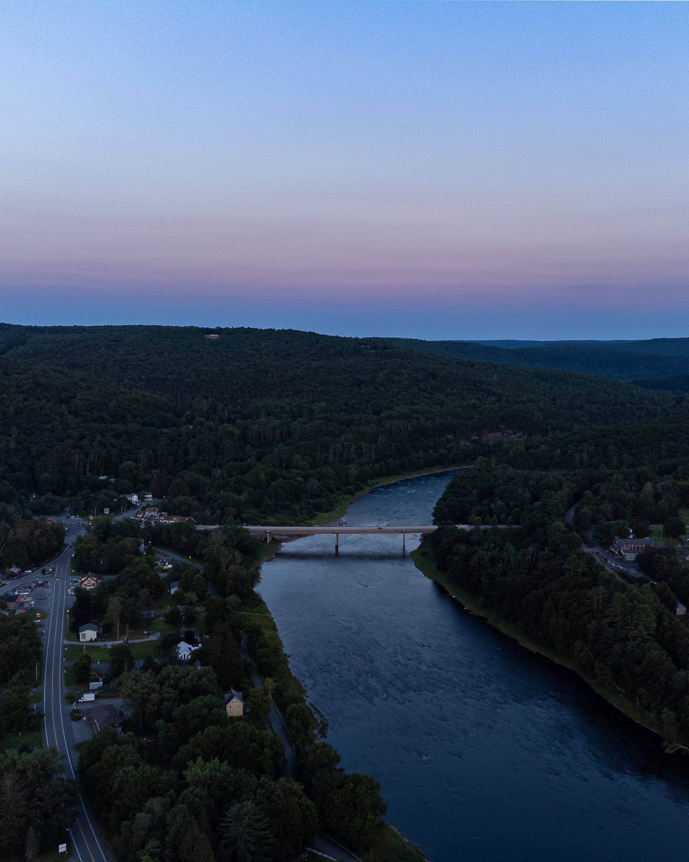 Aerial view of a river flowing through a valley with a bridge crossing over it, surrounded by dense green forests and a small town with roads and houses, under a pastel-colored sky at dusk.