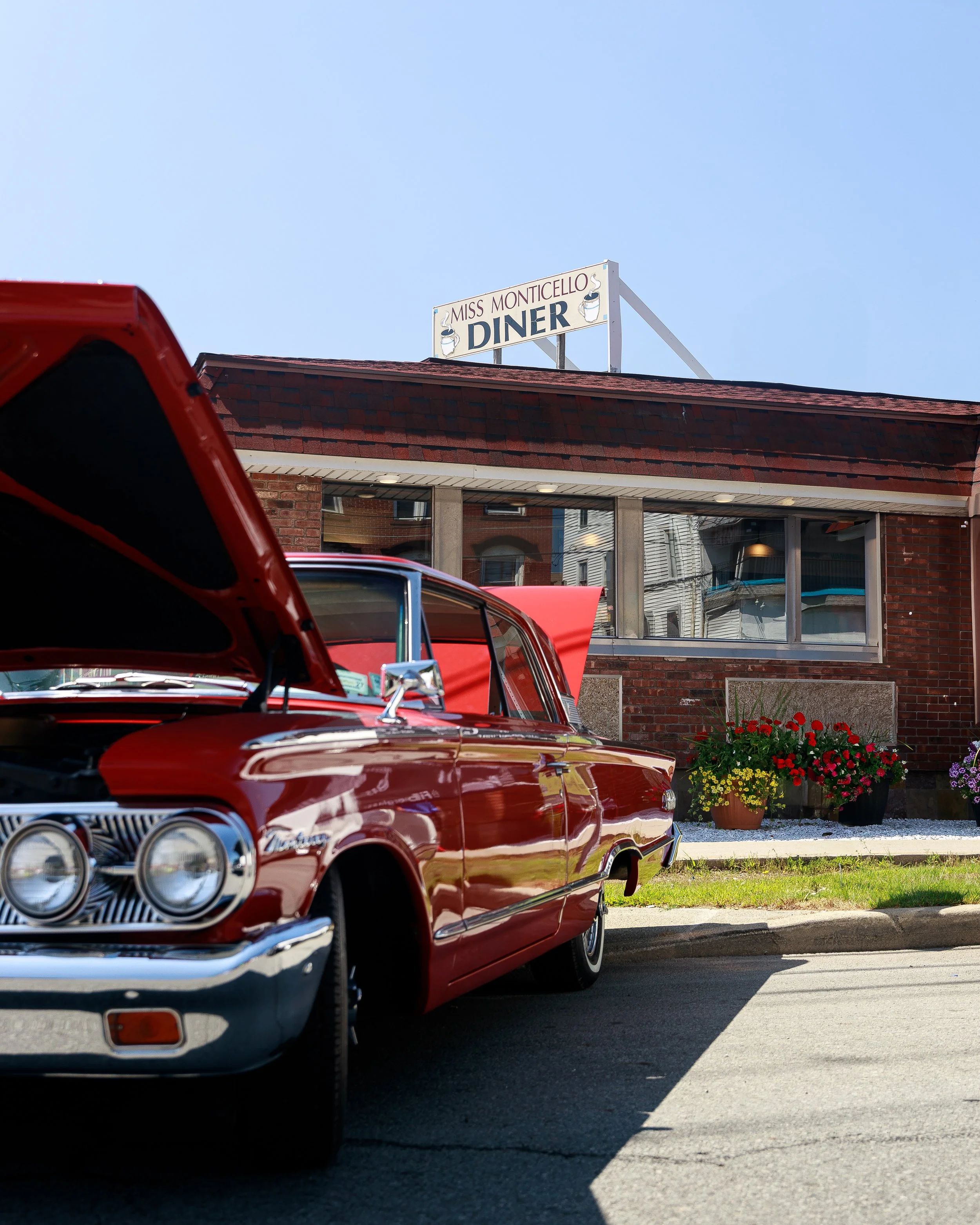 A classic red car parked in front of a brick diner with a sign that reads "Miss Monticello Diner." The car's hood is open, and there are colorful flowers in pots outside the diner.