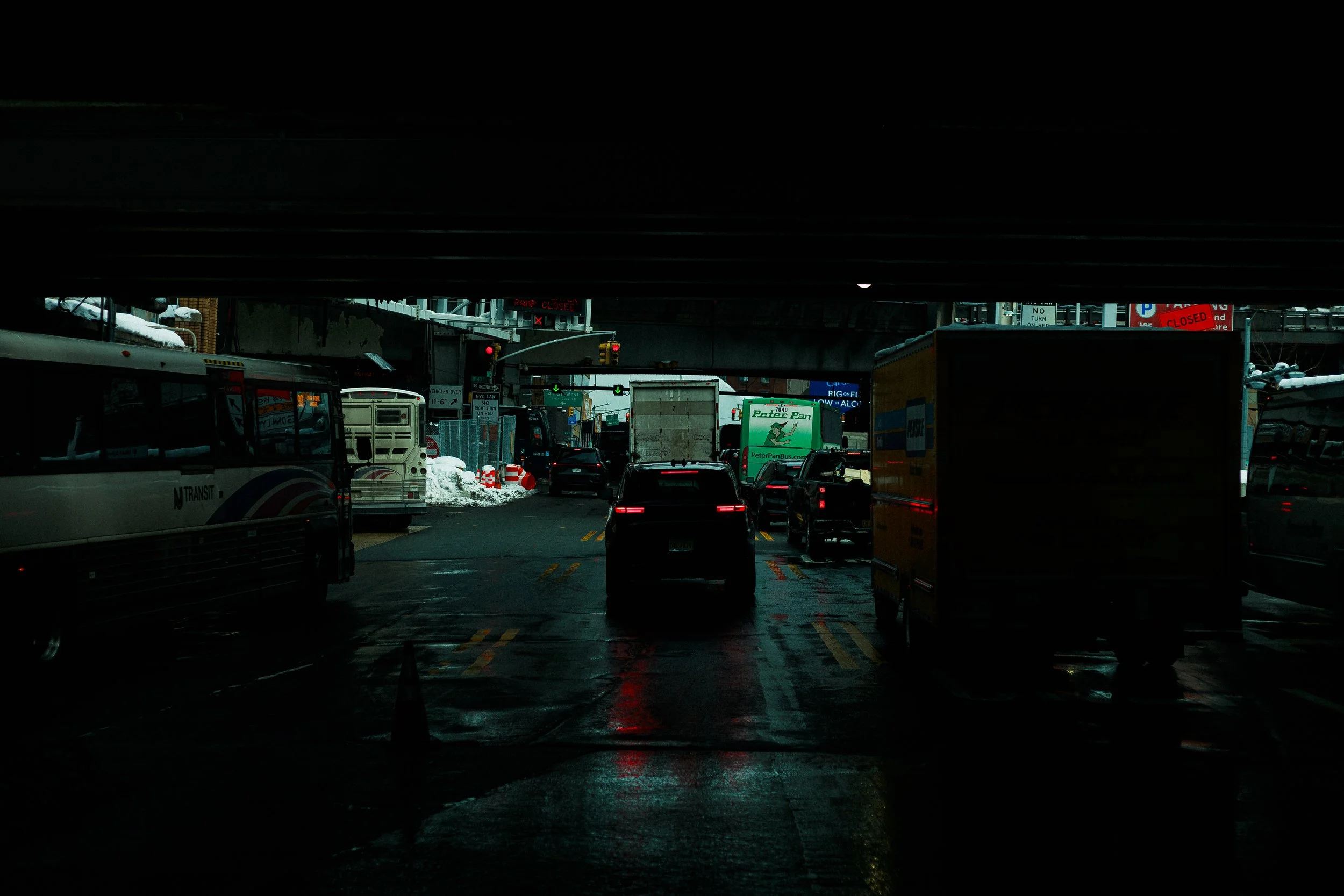 City street scene under a bridge, cars and buses in traffic, wet pavement, snow on the sides, and traffic lights.