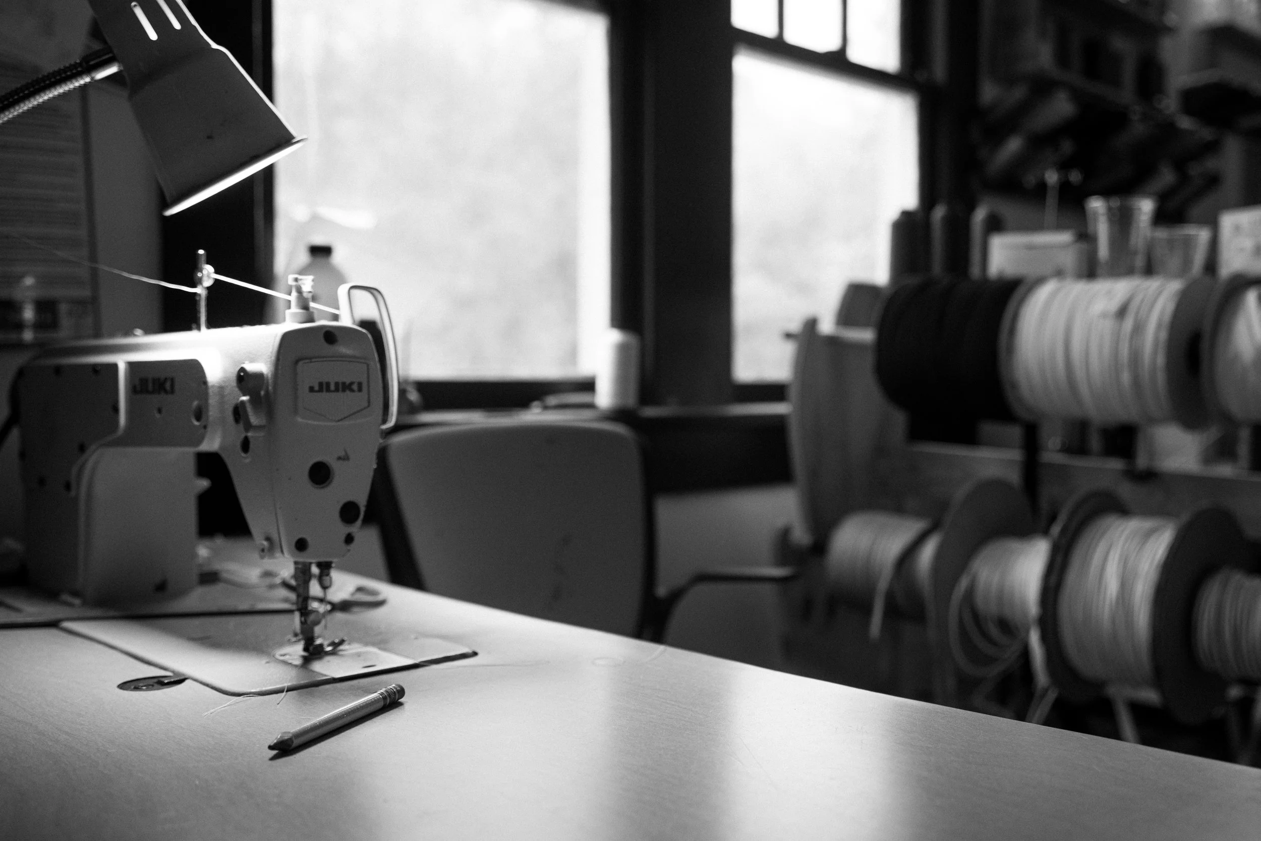 A sewing machine on a work table with spools of thread on shelves, illuminated by a desk lamp, in a craft room or sewing workshop.