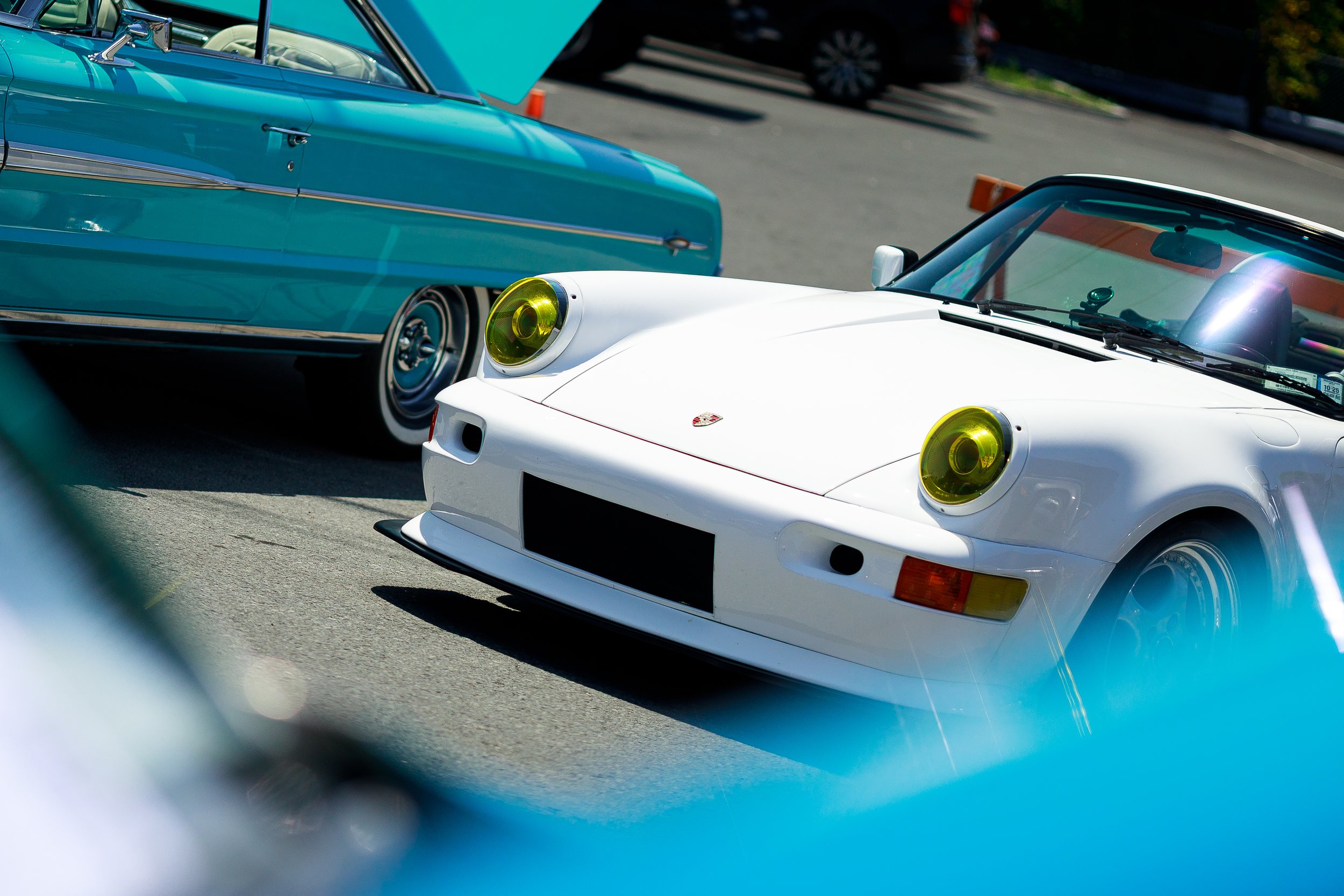 A white vintage Porsche sports car with yellow-tinted headlights parked among other classic cars, including a light blue car, at a car show or gathering.
