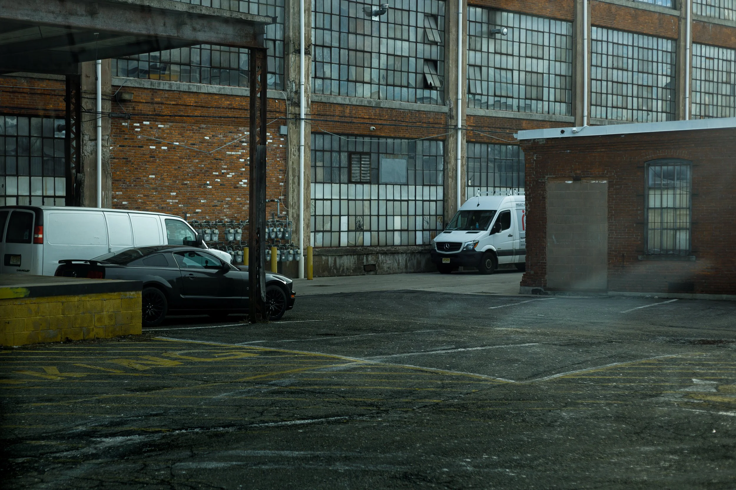 An urban alleyway scene with parked vehicles, including a black sports car, a white van, and a delivery van, against an industrial brick building with large windows and utility meters.