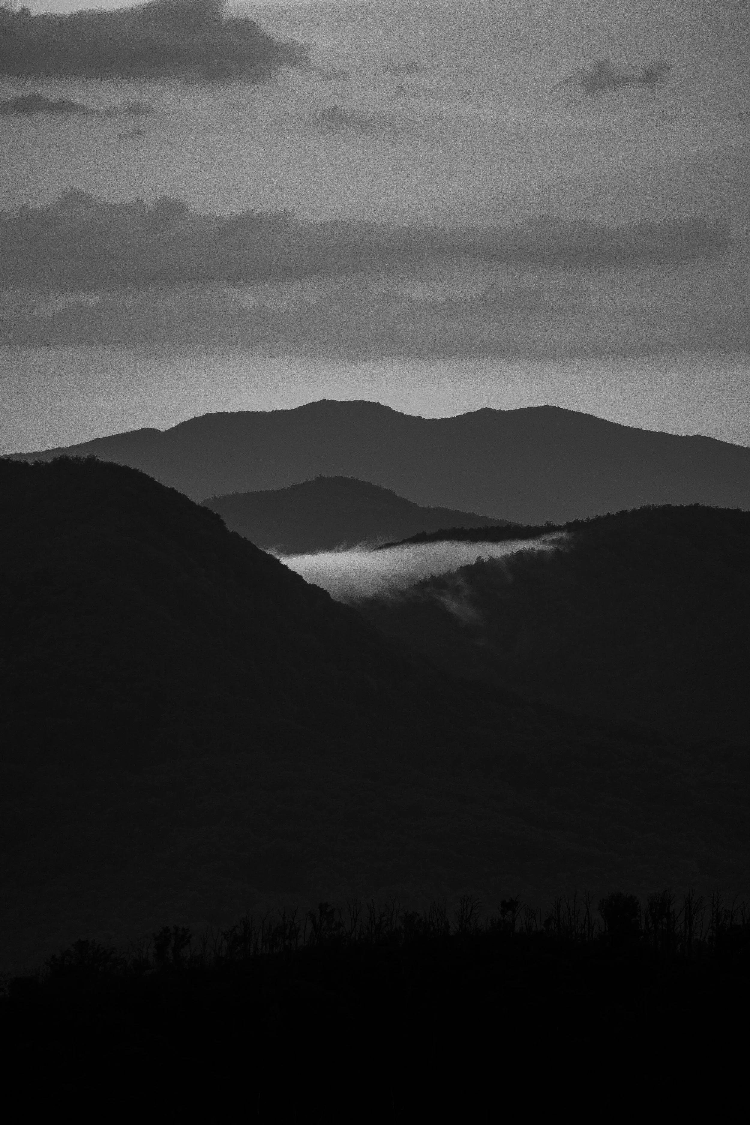 Black-and-white photograph of a mountain landscape with multiple layers of ridges and a few clouds in the sky.