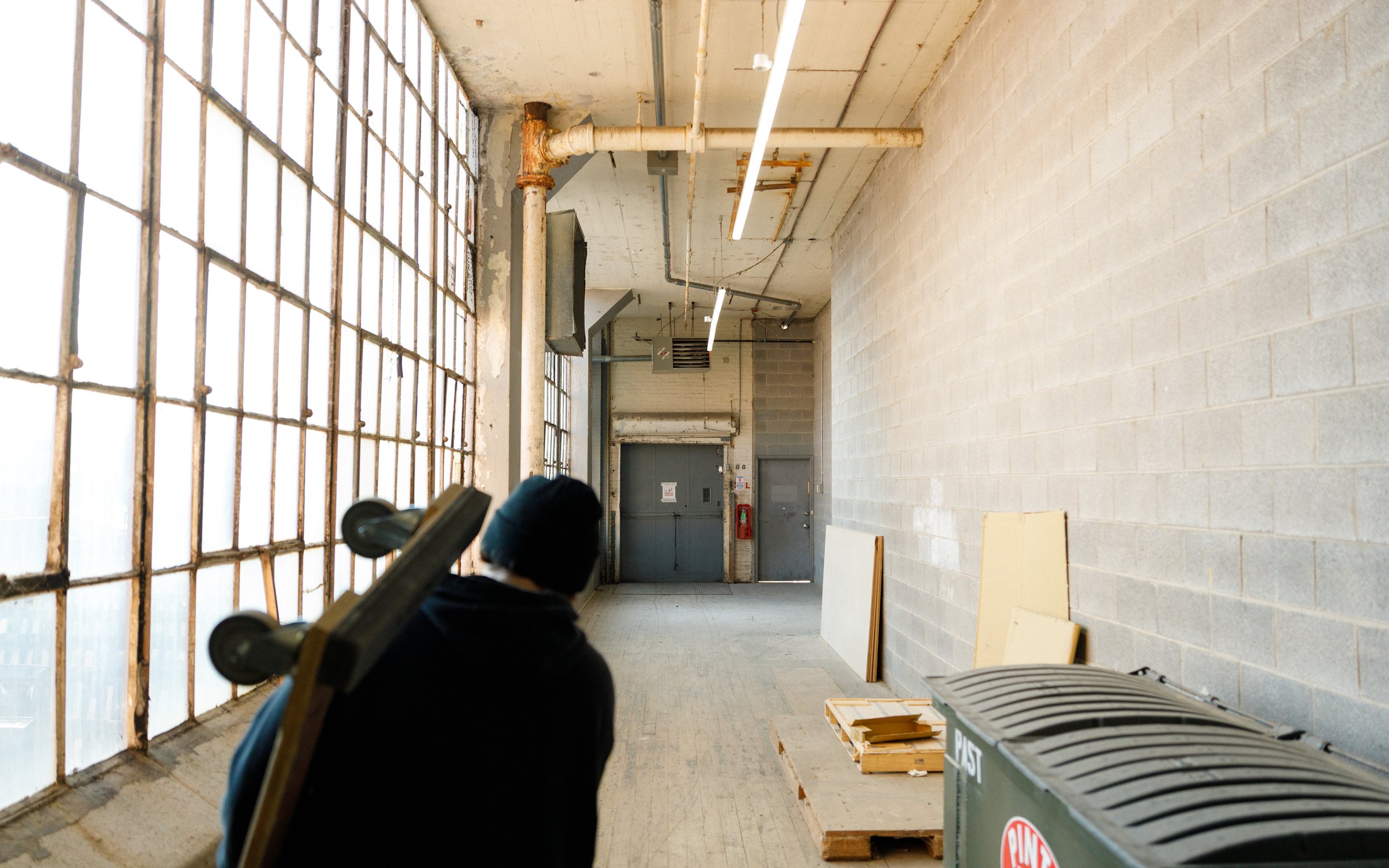 A person with a skateboard strapped to their back is walking through an industrial-style corridor with large, grid-patterned windows on the left, and a cinder block wall on the right. The corridor has a high ceiling with exposed pipes and fluorescent lighting, and a green trash bin is visible in the foreground.