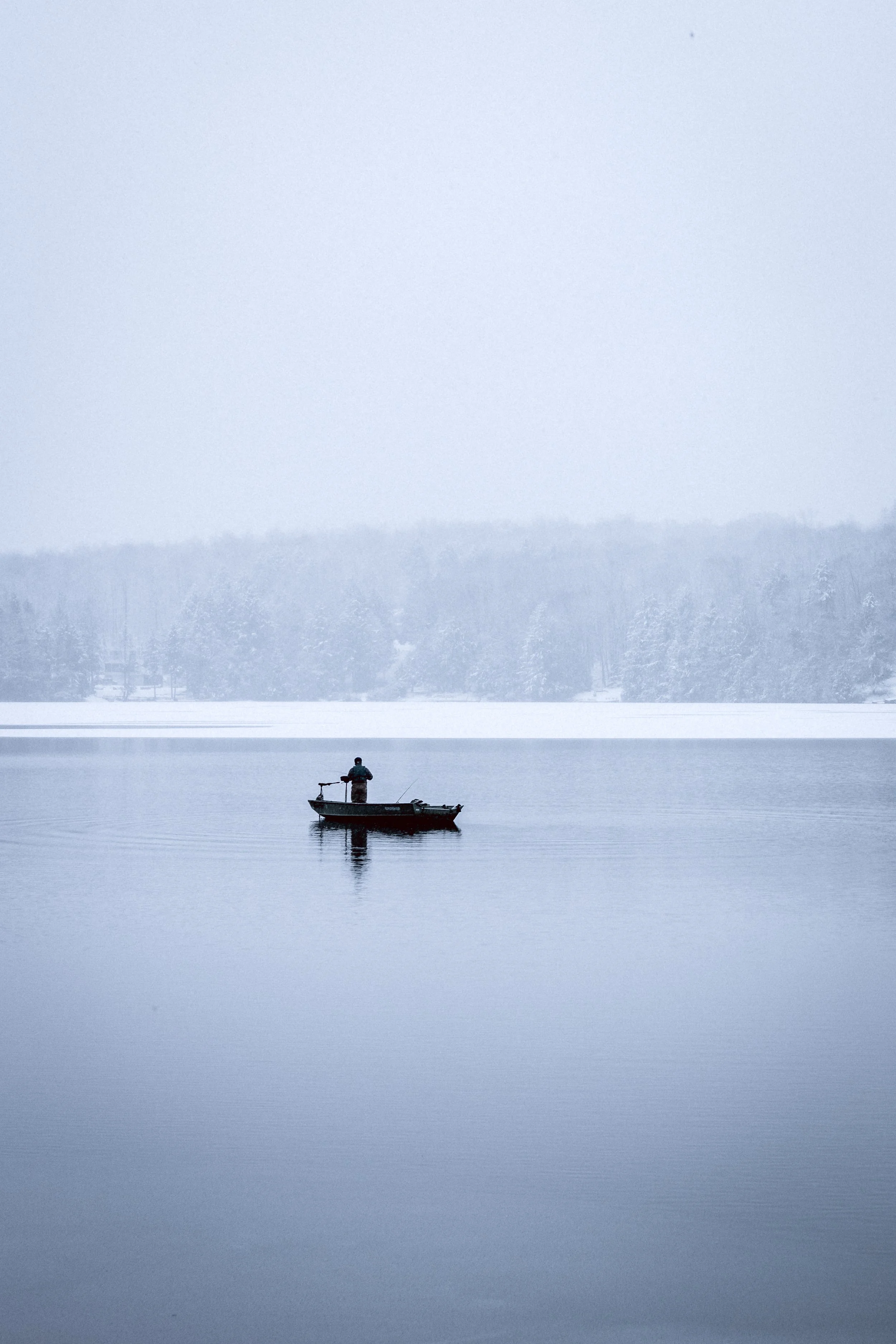A person fishing from a small boat on a calm, icy lake during winter, with snow-covered trees in the background.