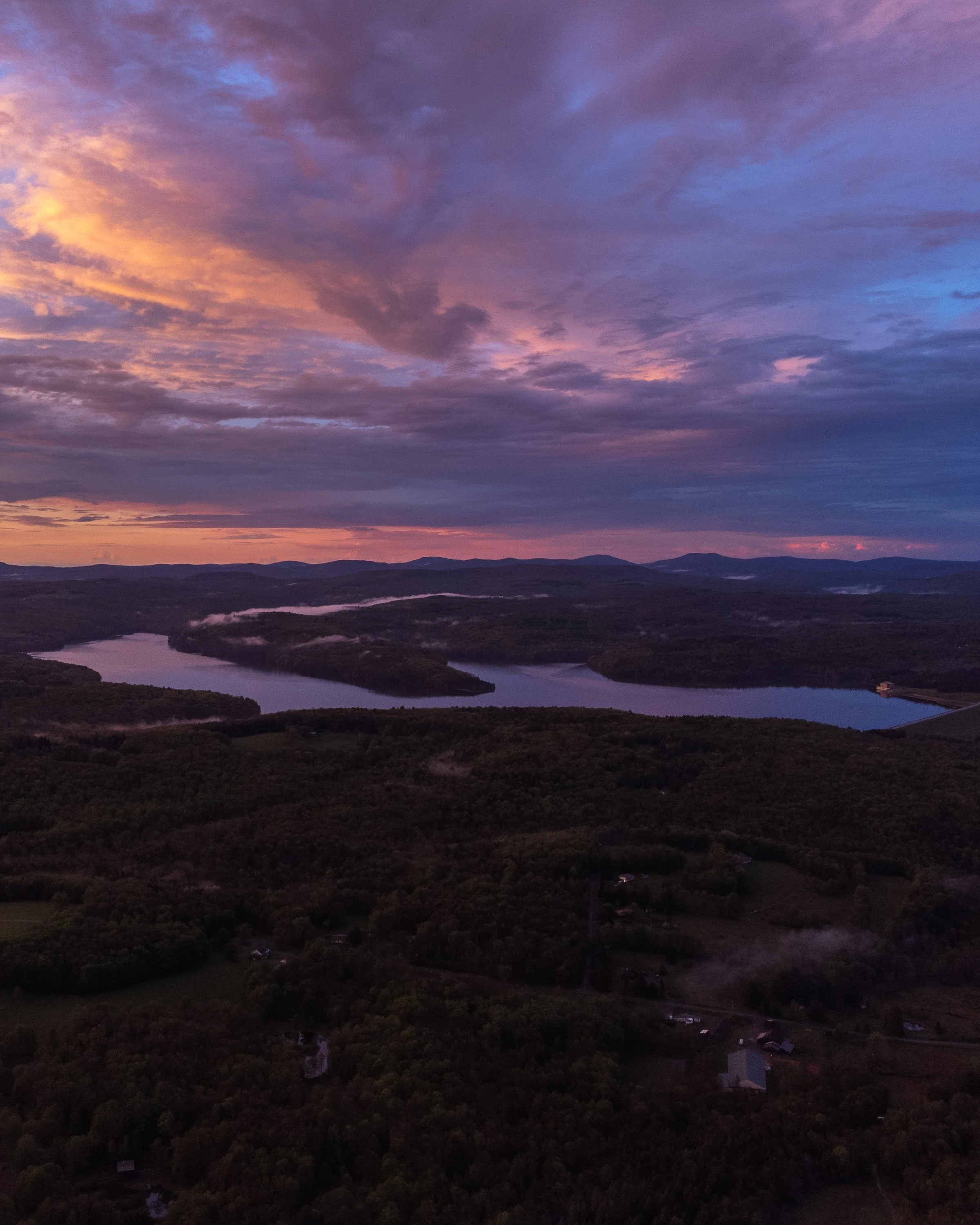 Aerial view of a landscape during sunset, featuring a winding river, rolling hills, and a colorful sky with clouds.