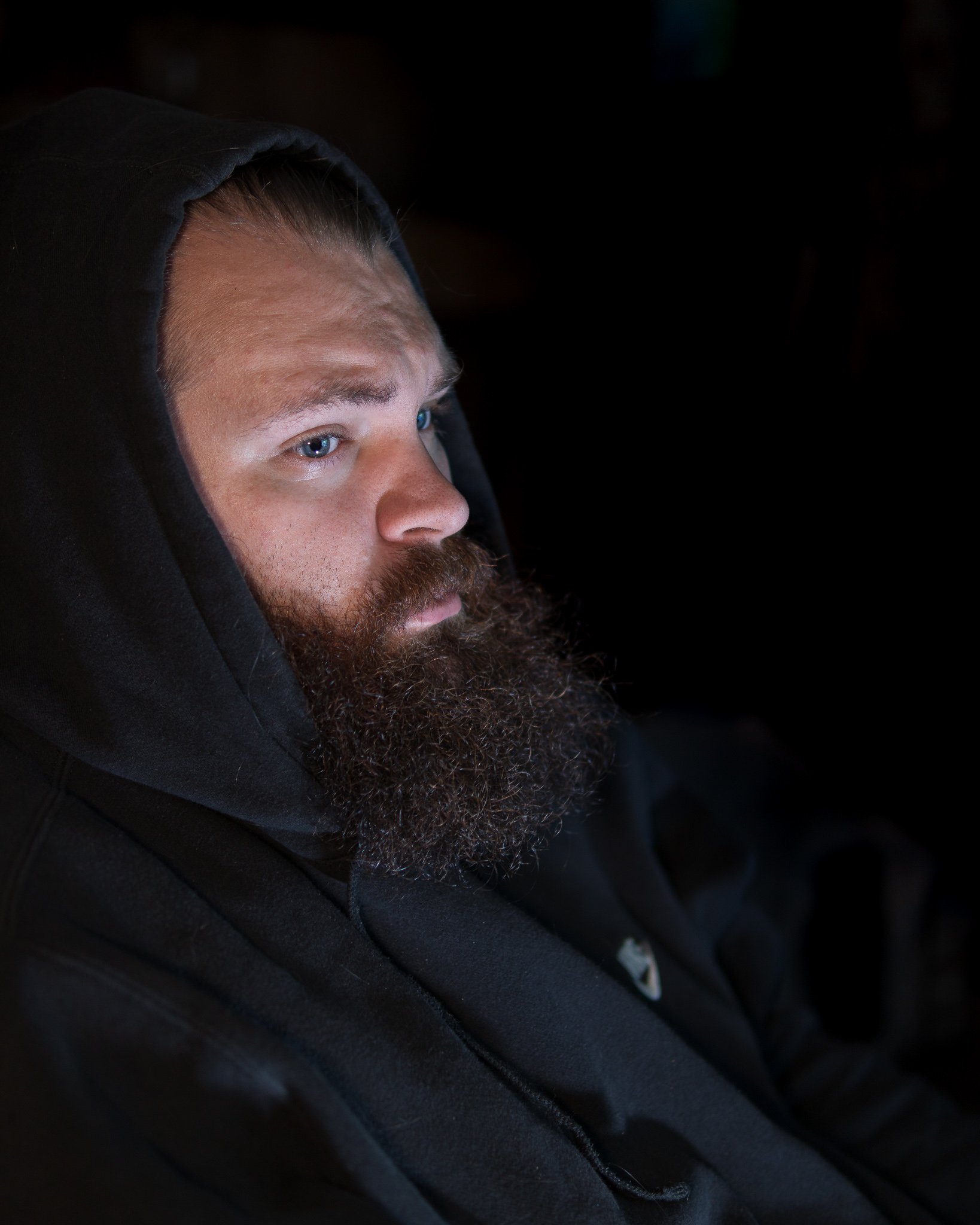 A man with a beard and blue eyes wearing a black hoodie, looking intently to the side in a dark setting.