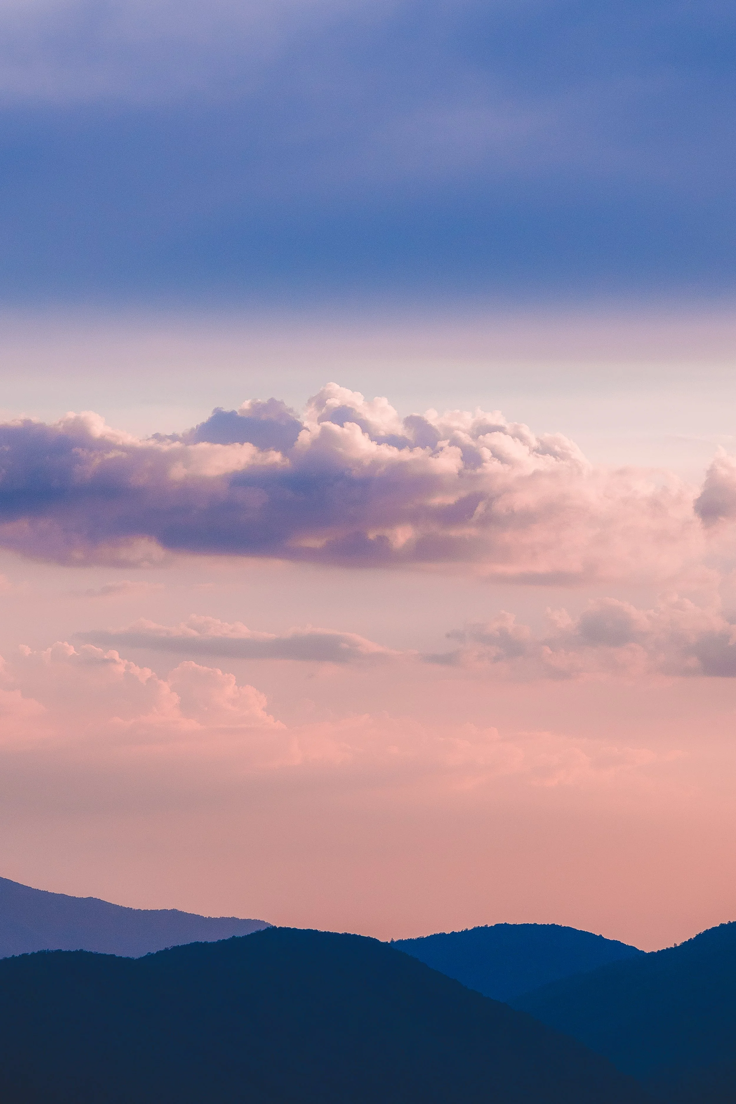 A mountain range with layered dark silhouettes under a pink and purple sky with fluffy clouds at sunset or sunrise.