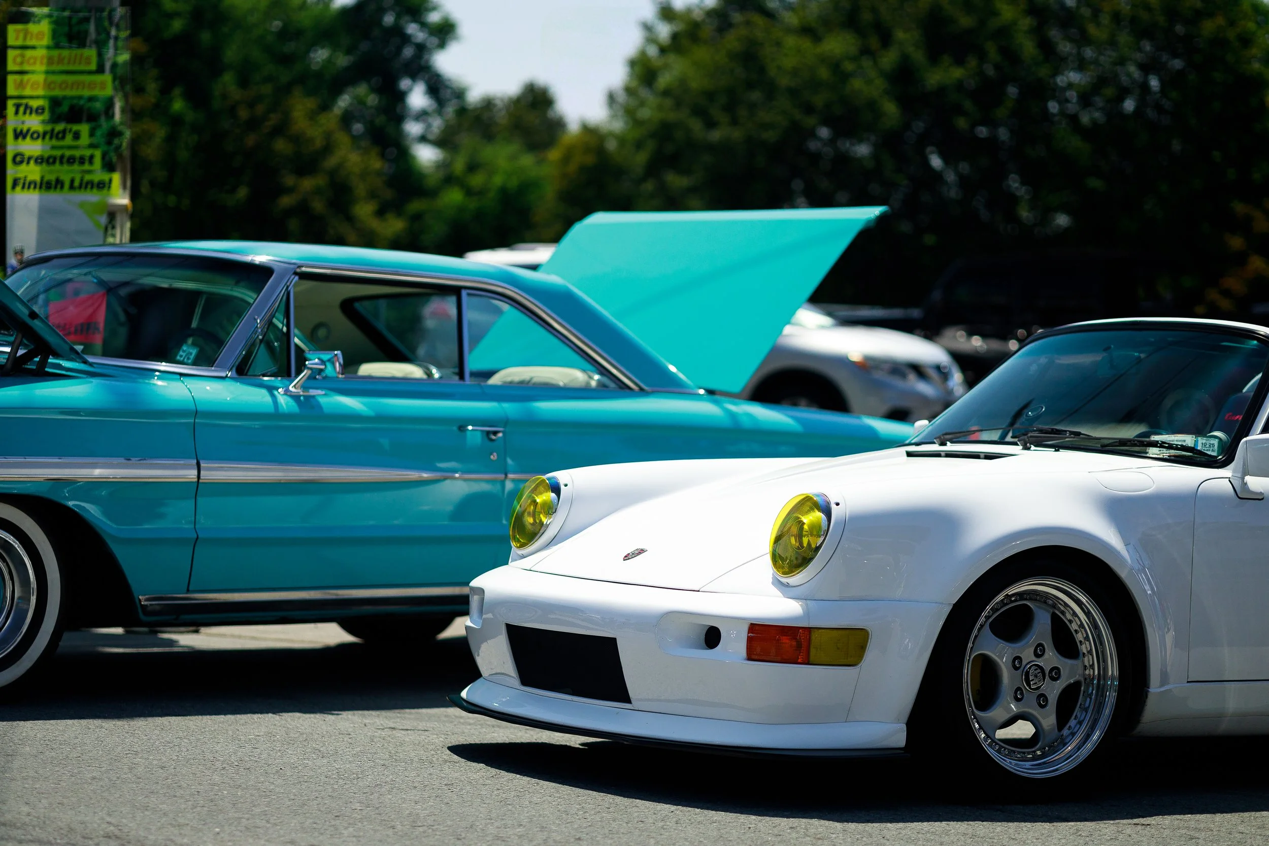 A white sports car with yellow headlights and a blue vintage car with open hood parked at a car show, with trees in the background.