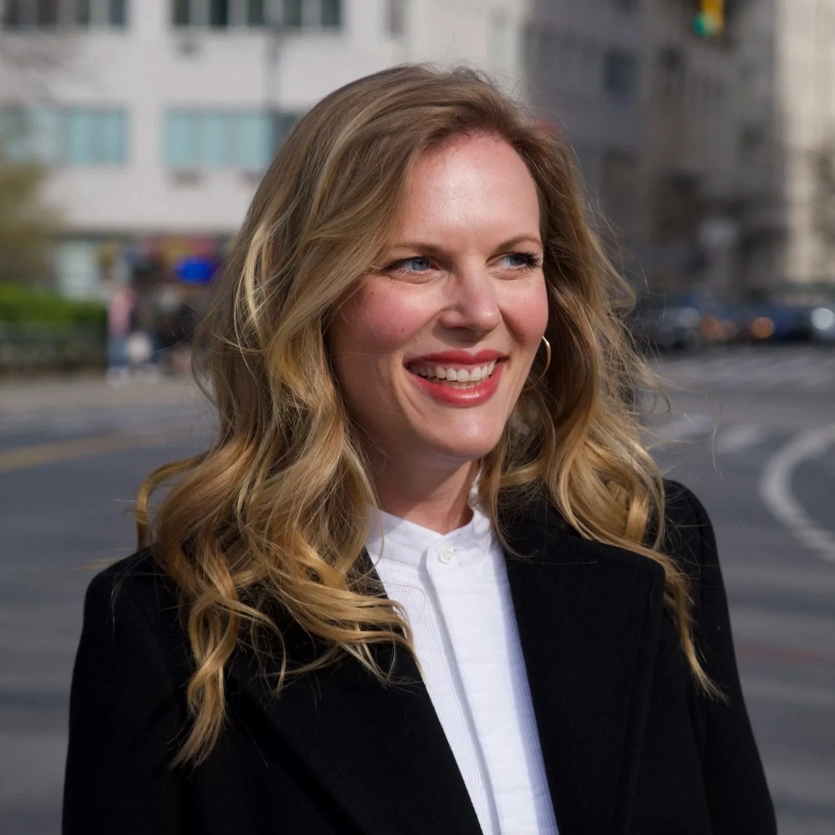 Headshot of Victoria Brooks. Blonde woman, smiling, looking off into the distance. Busy city background.