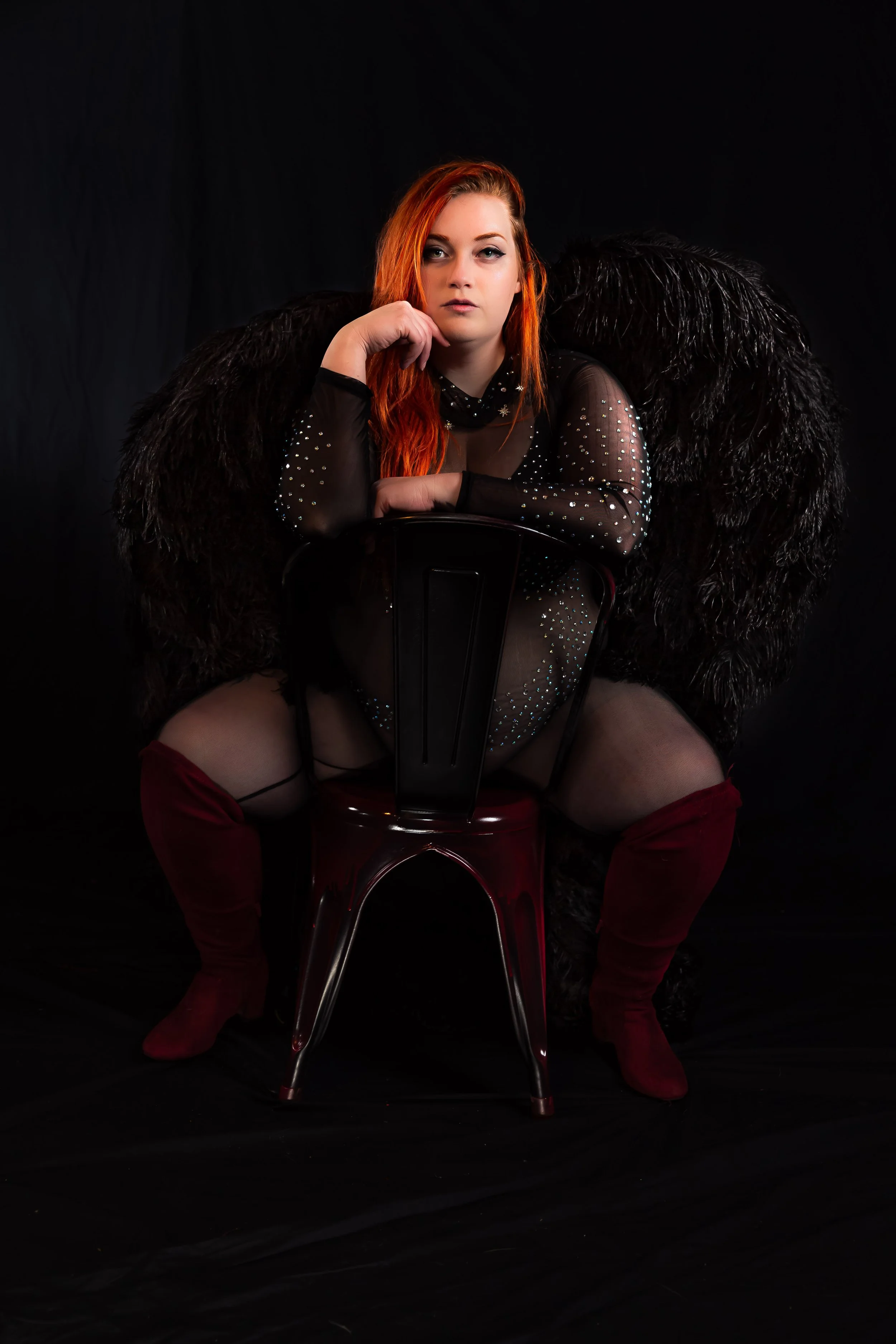 Woman with copper hair in black lingerie and thigh-high boots seated on a chair in a Maine boudoir studio