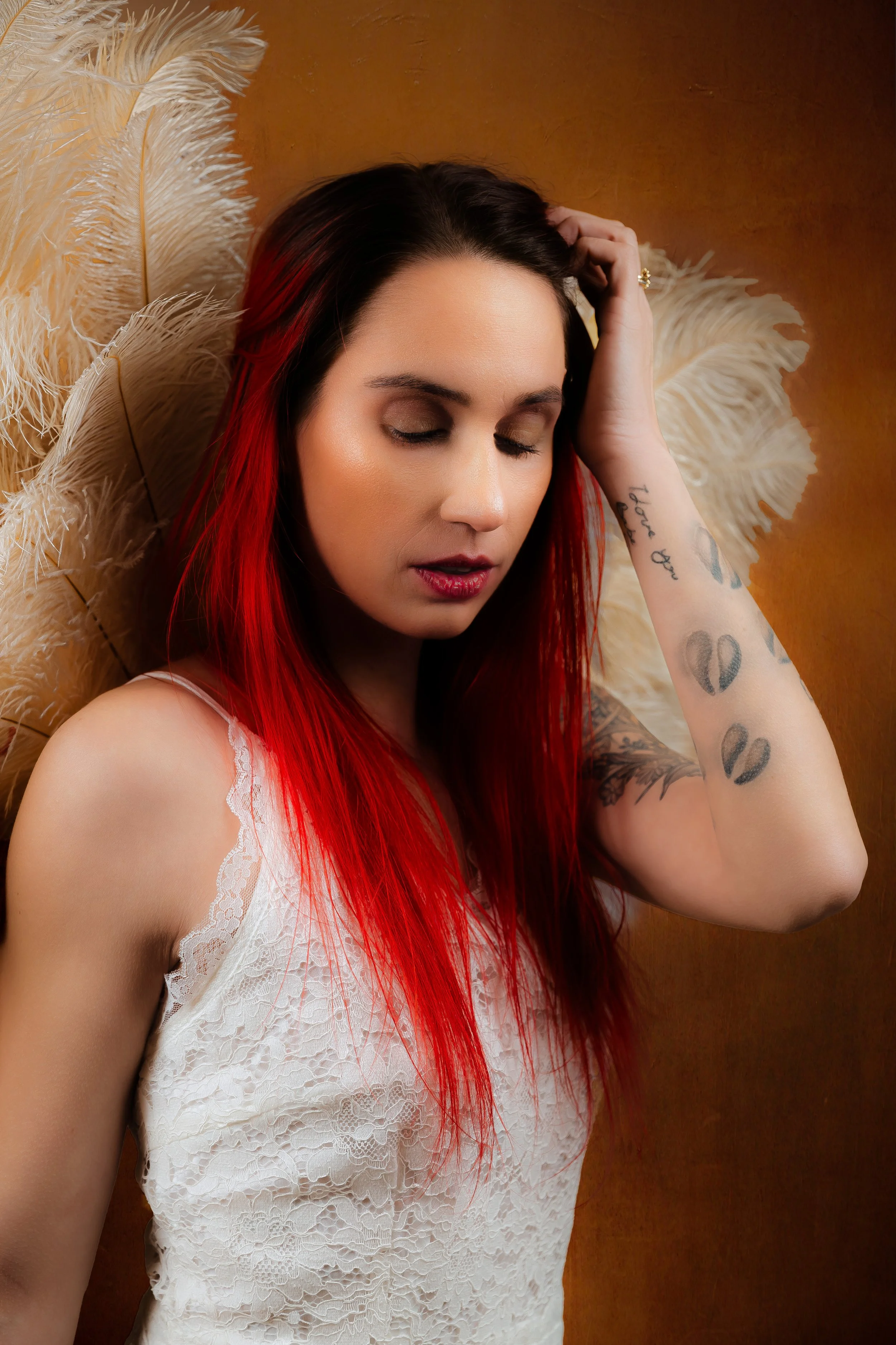 Woman with red hair in a white lace top posing in front of feather fans in a Maine boudoir studio