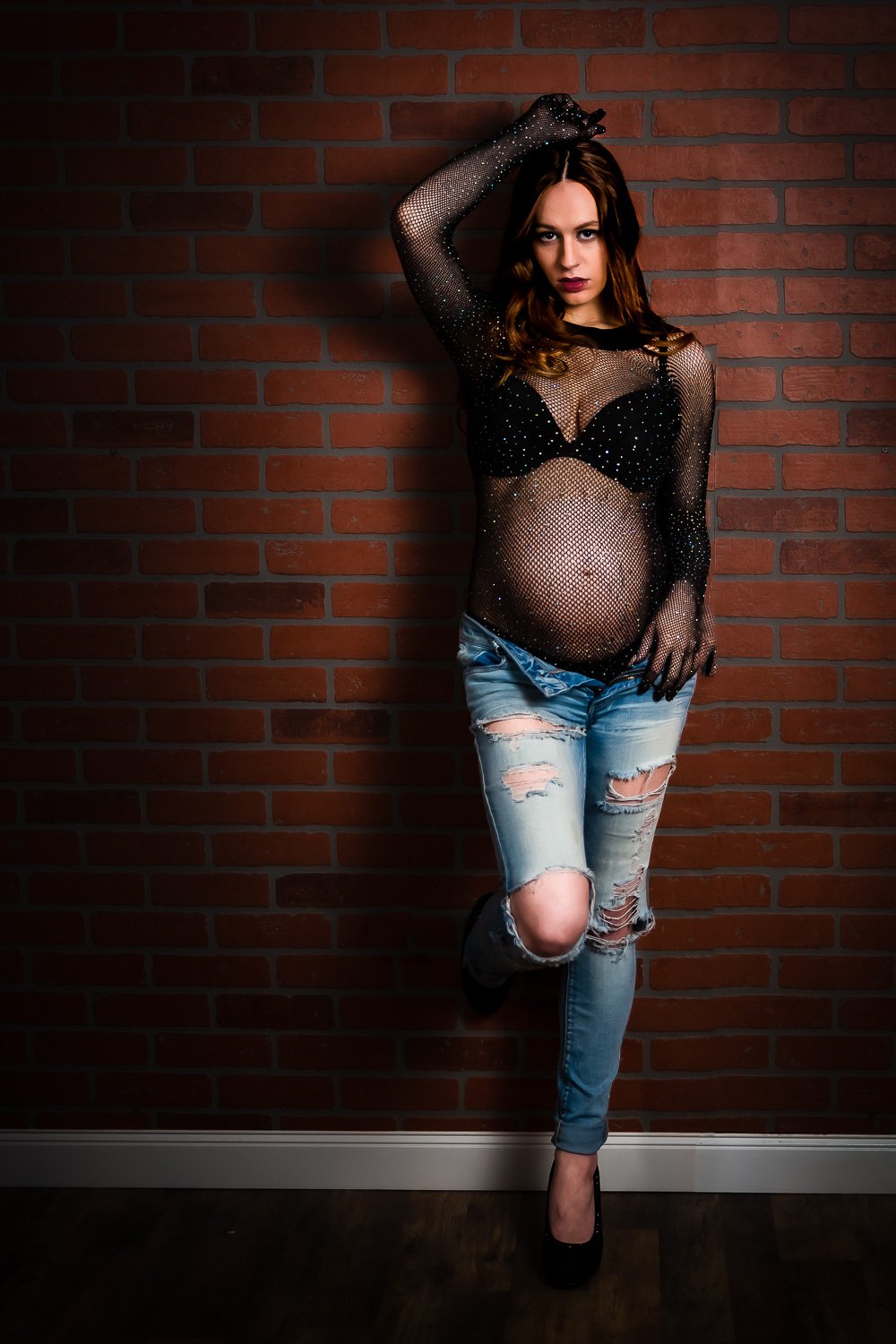 Expecting mother in a sheer black top and jeans against a brick wall during a maternity boudoir session in Maine