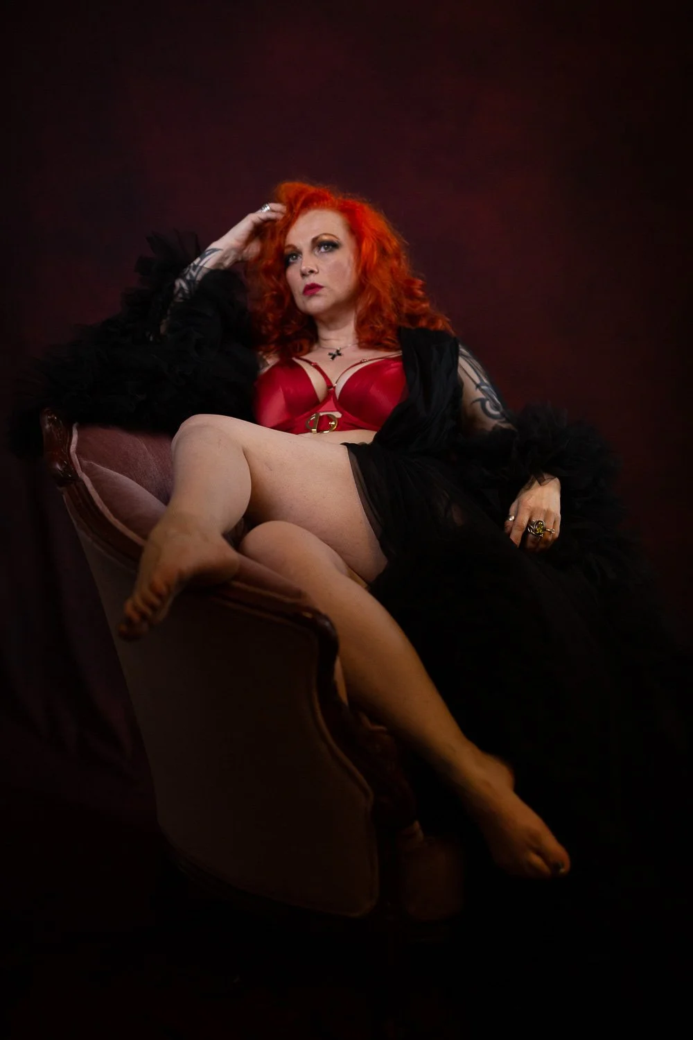 Red-haired woman in red lingerie and a black robe seated in a dark chair during a boudoir session in Bangor, Maine