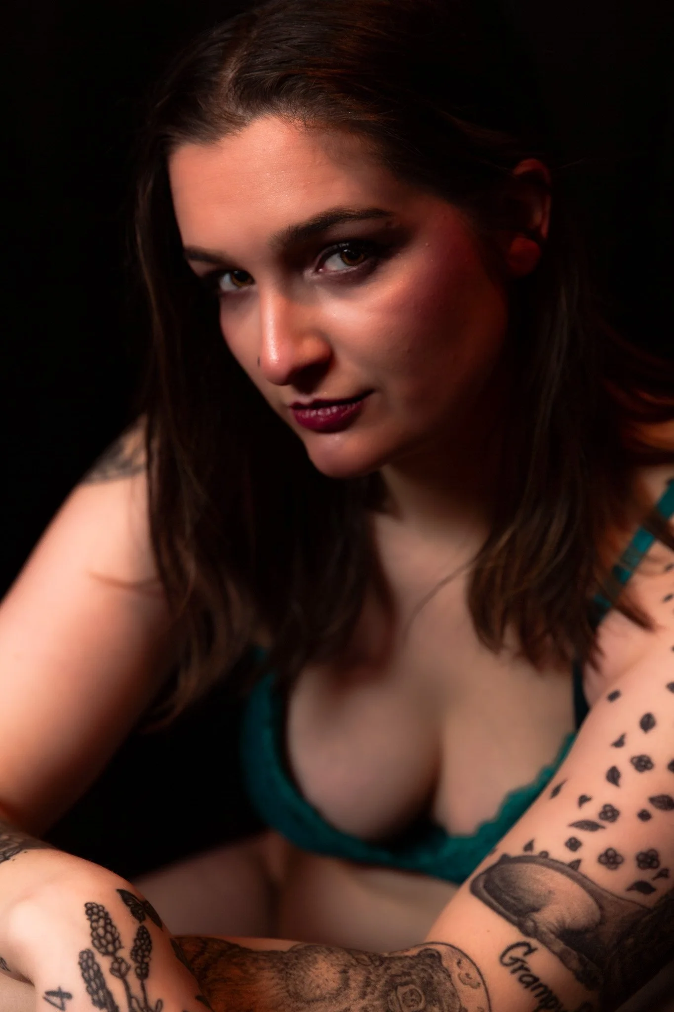 Tattooed woman in a teal bra leaning toward the camera during a boudoir session in Maine