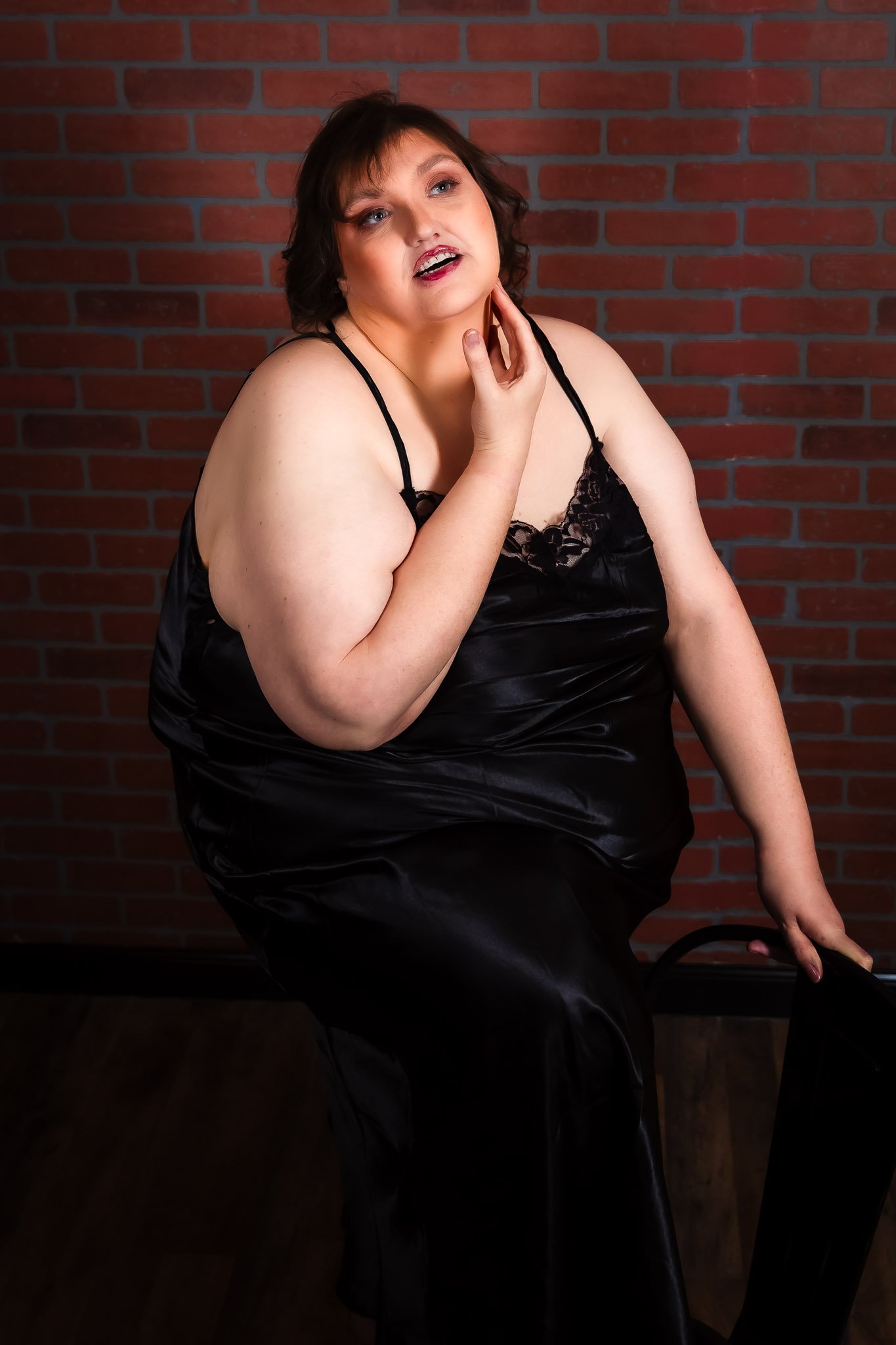 Curvy woman in a black satin slip seated against a brick wall during a boudoir session in Maine