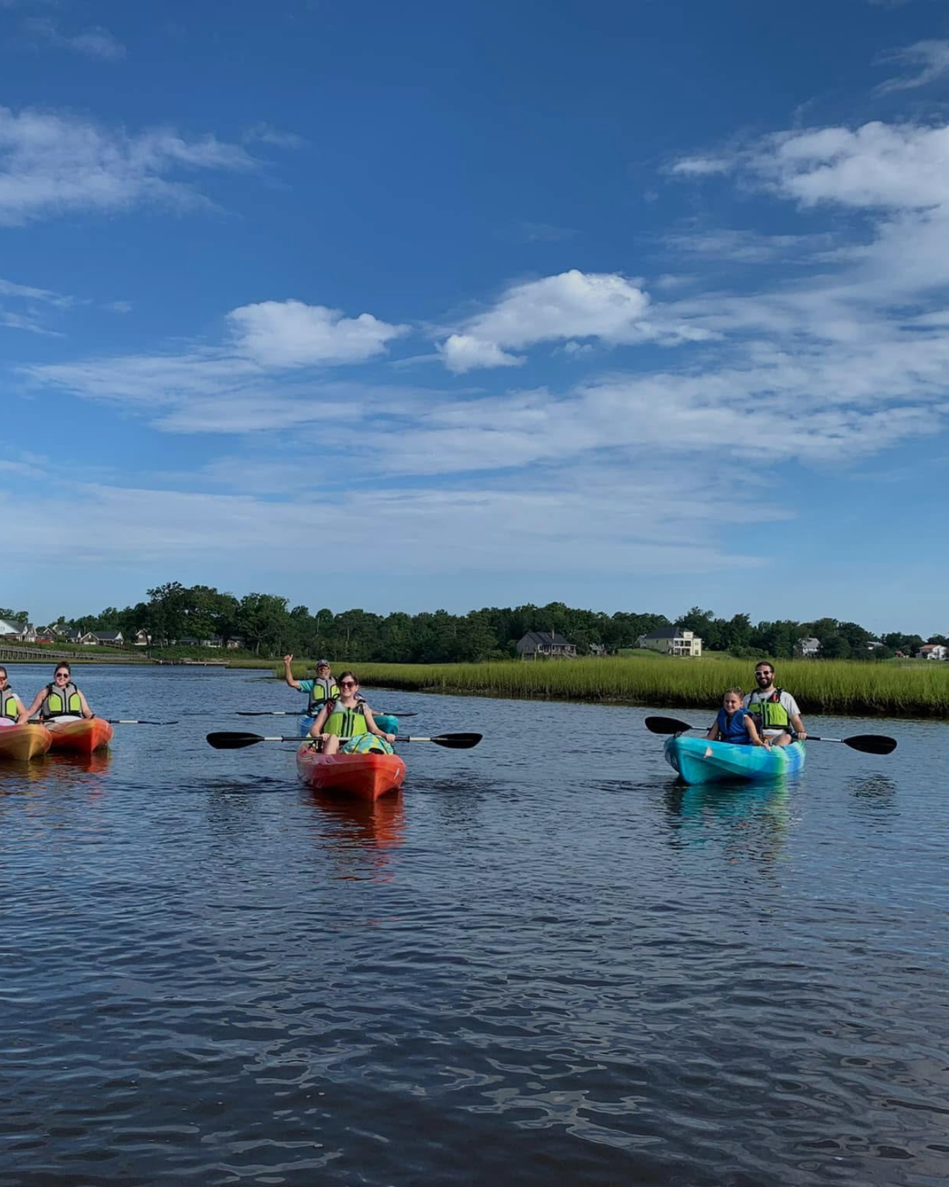 Family kayaking on a calm water body under cloudy sky, with two kayaks in the foreground and a person kayaking in the background.