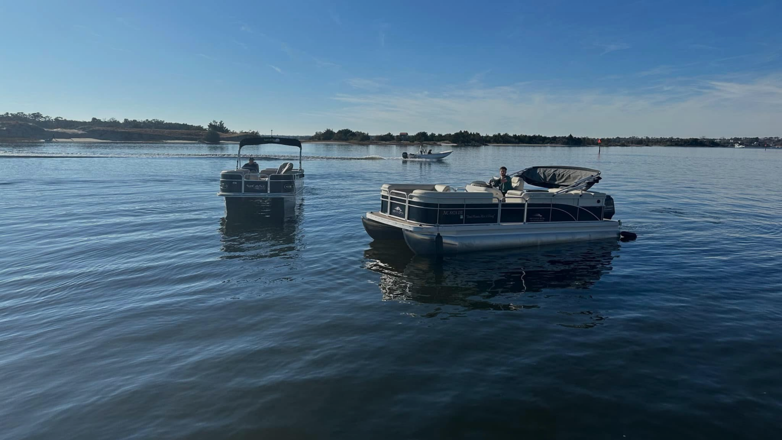 A group of people on a pontoon boat by the water, with blue sky and some clouds, and a shoreline in the background.